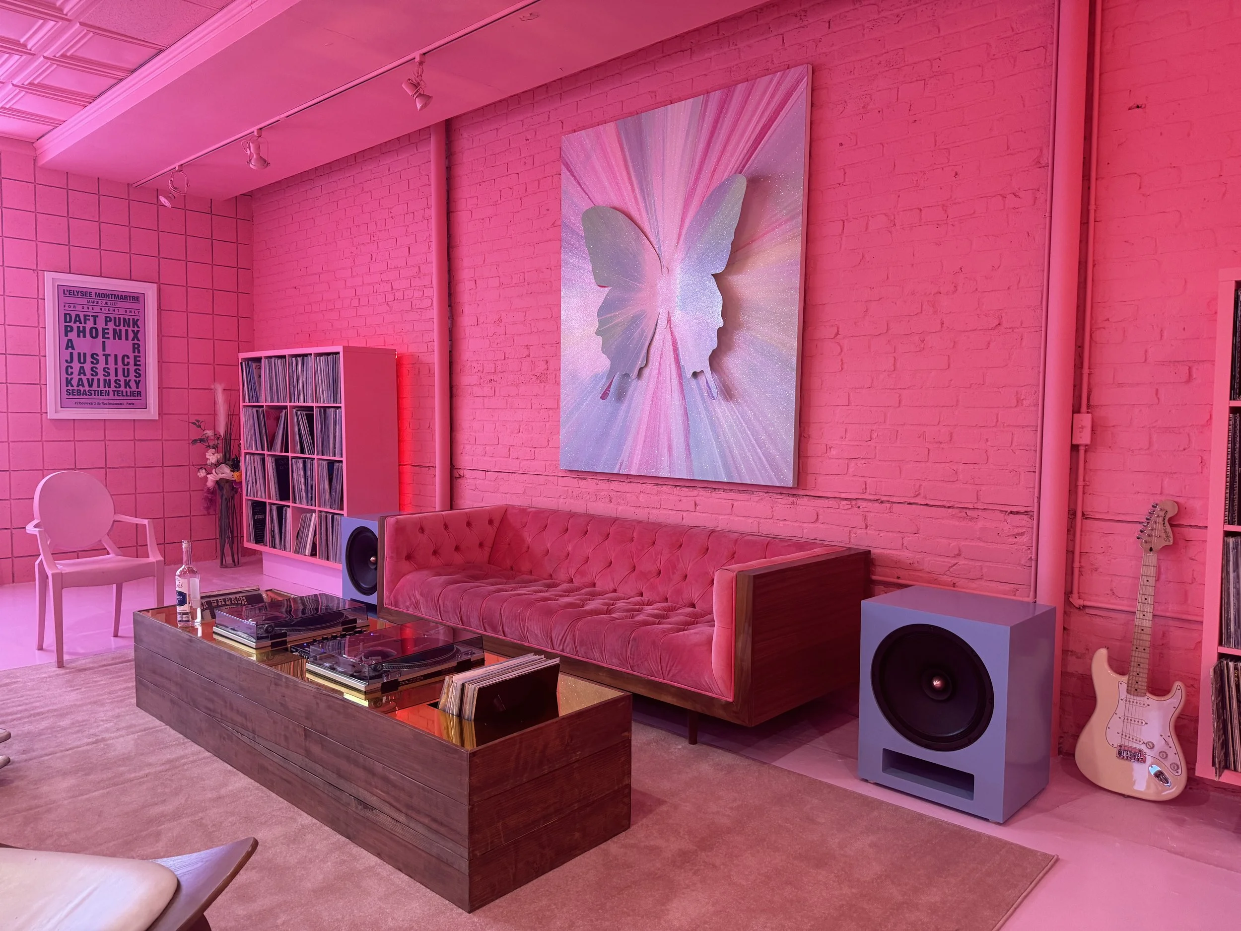 Room with pink lighting featuring a pink tufted sofa, butterfly art on the wall, vinyl records on wooden shelves, a large speaker, a guitar, and a framed poster. A glass-top coffee table with turntables and records is centered on a rug.