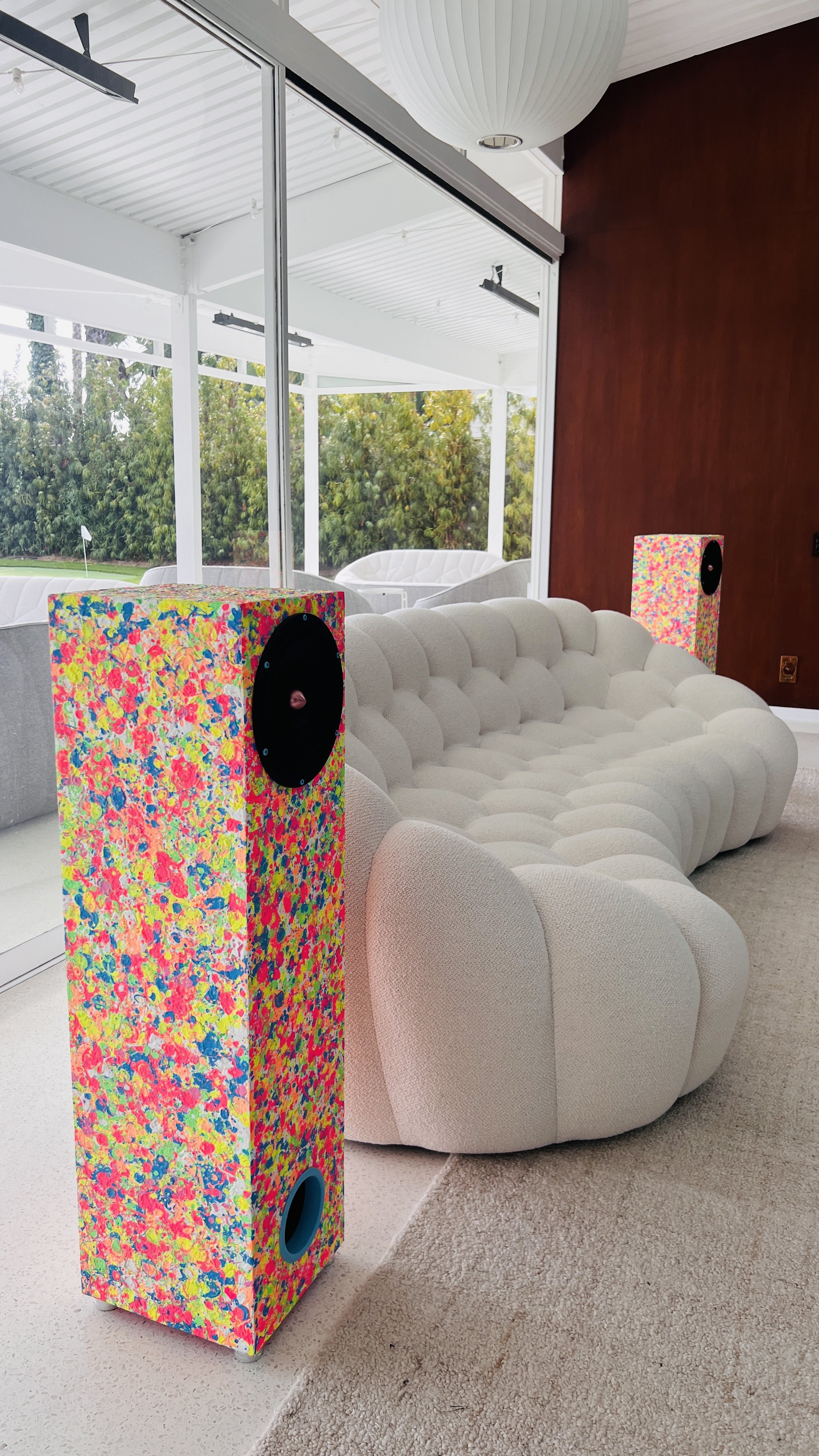 A modern room featuring a white, tufted sofa and two speakers with colorful, abstract designs. There's a large glass window, a white pendant light, and visible greenery outside.