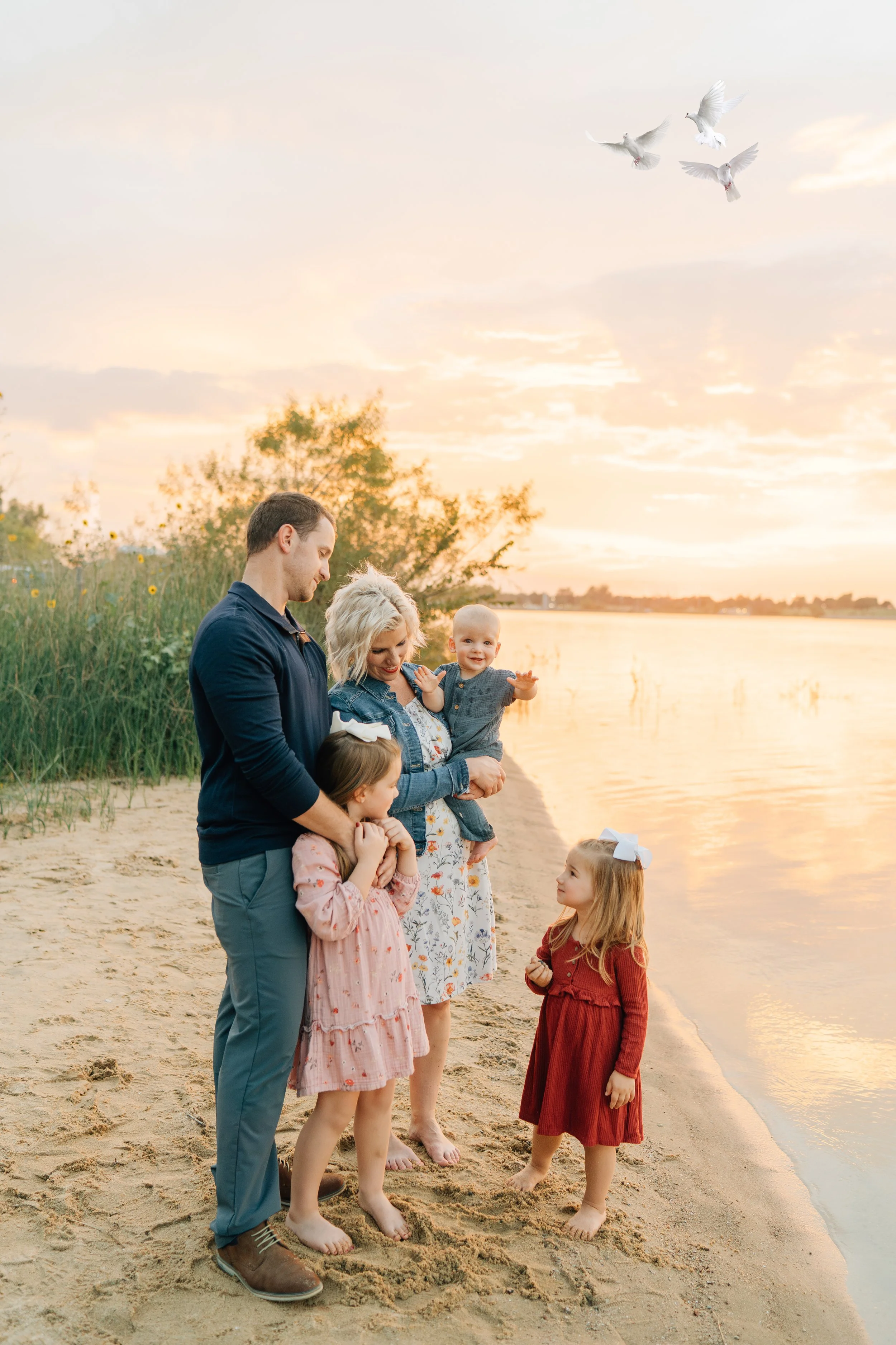 Family of five, including two adults and three children, on a sandy beach during sunset, with four white doves flying overhead.