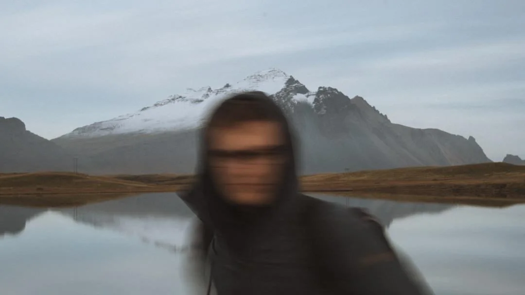 blurry image of moutain landscape in background and man with hoodie and glasses in foreground