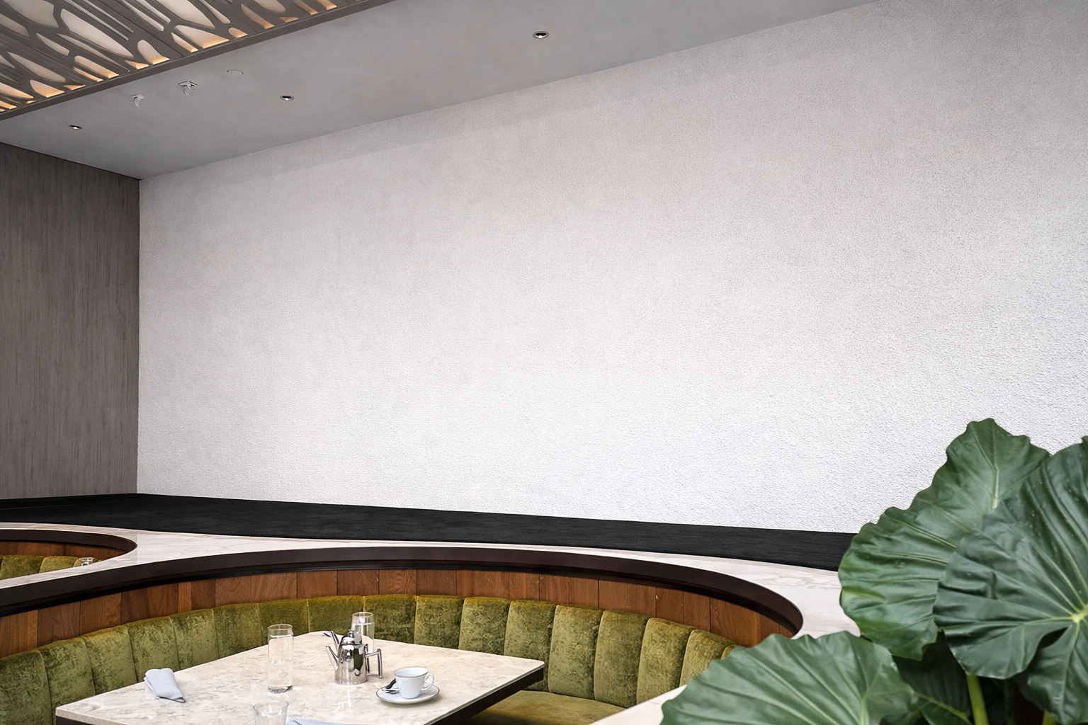 Empty restaurant corner with a marble table set with glasses, a coffee cup, and a teapot, surrounded by a green velvet cushioned banquette, with a large leafy plant in the foreground and a white textured wall in the background.