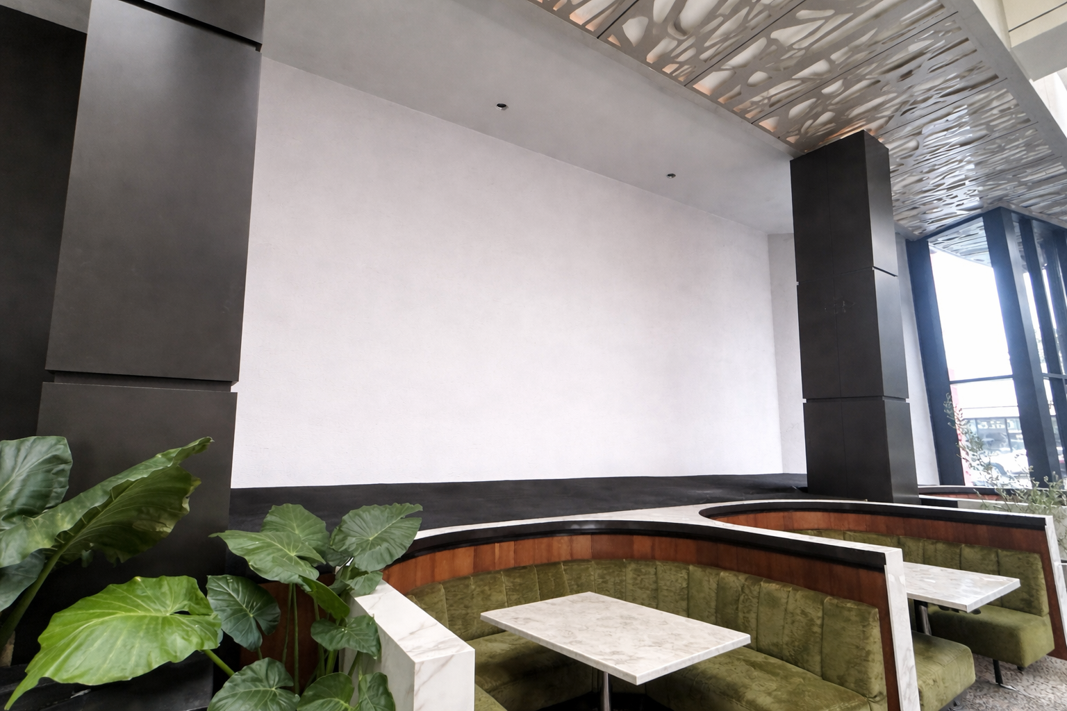 Interior of a modern space with a curved green velvet booth with marble tables, large black pillar, tall plants, and large windows letting in natural light.