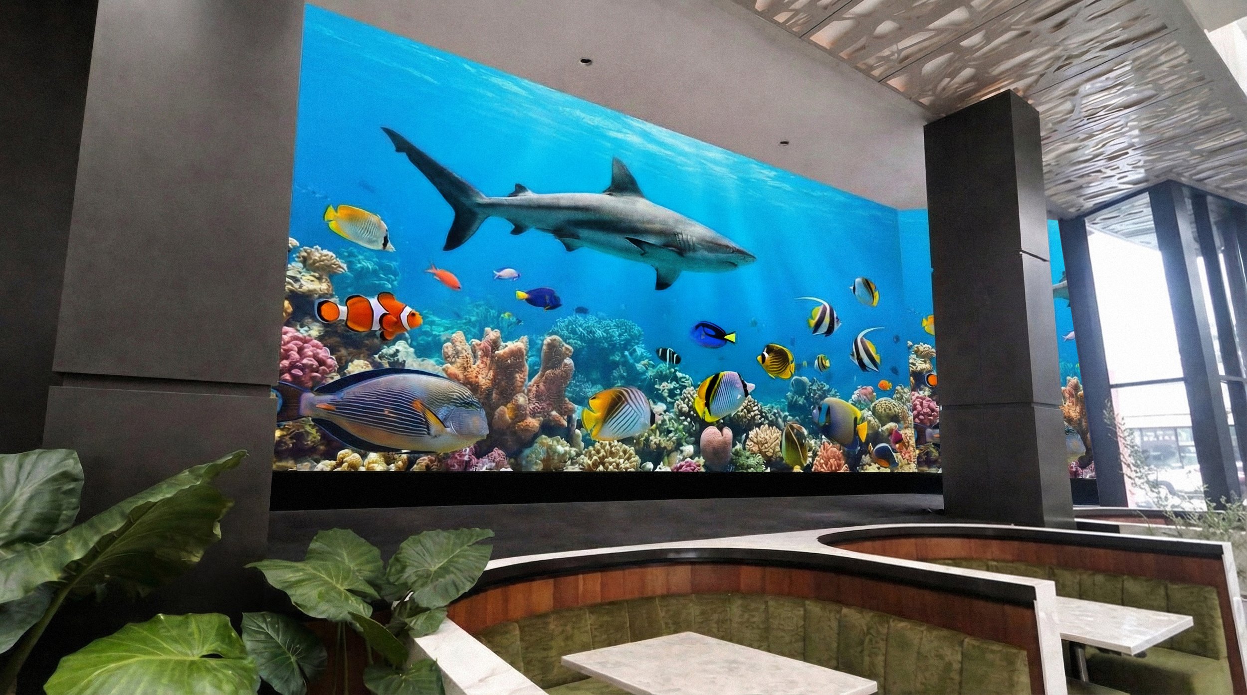 Indoor aquarium with a large wall display of colorful fish and a shark swimming above a coral reef, seating area in foreground with green plants.