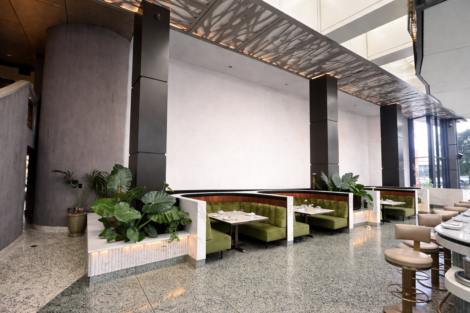 Interior of a modern restaurant with green cushioned booths, beige bar stools, potted plants, terrazzo flooring, and large glass windows allowing natural light.
