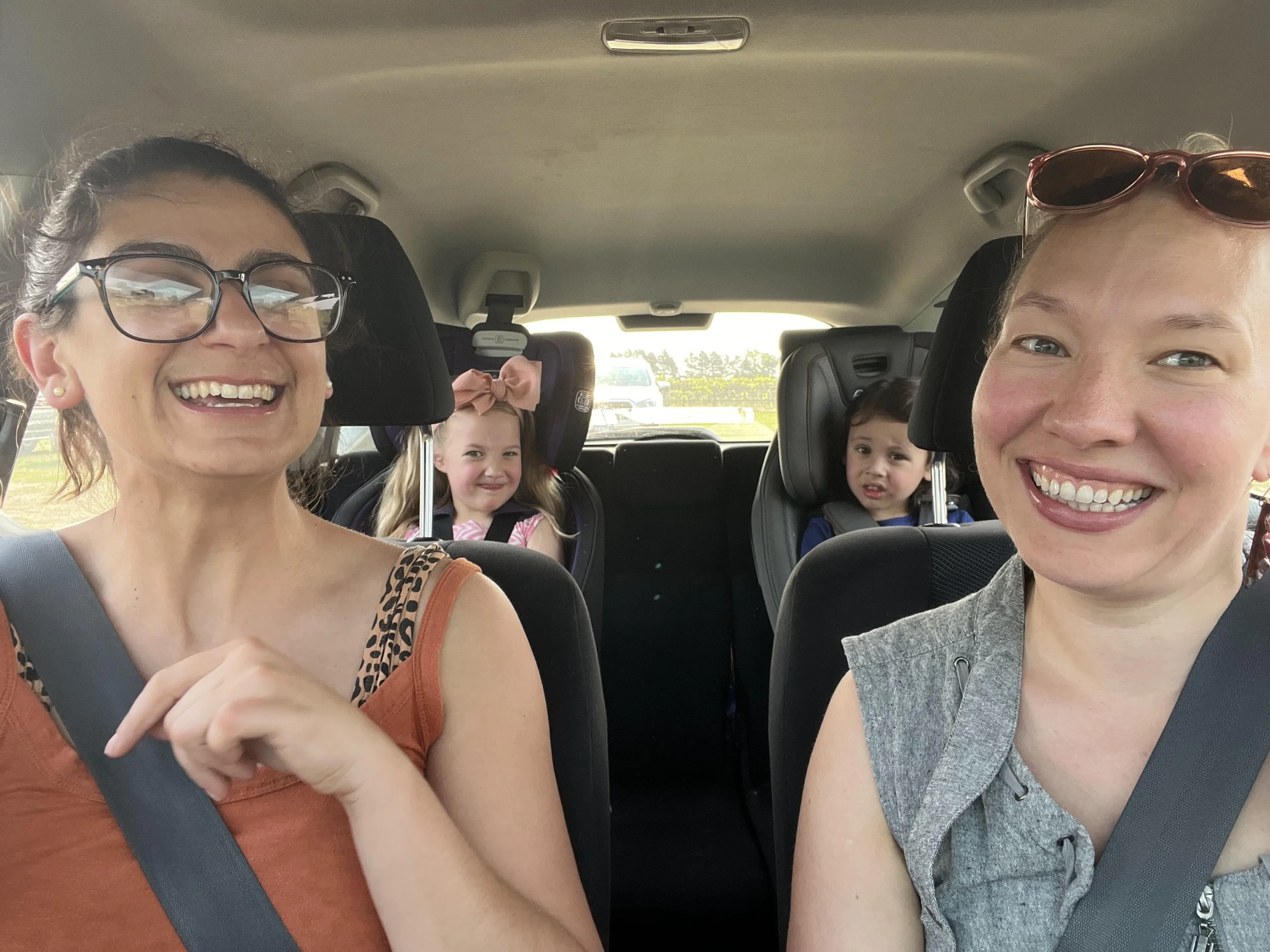 An Arab woman in glasses and a white woman with sunglasses are driving with two white children in carseats in the back seat.