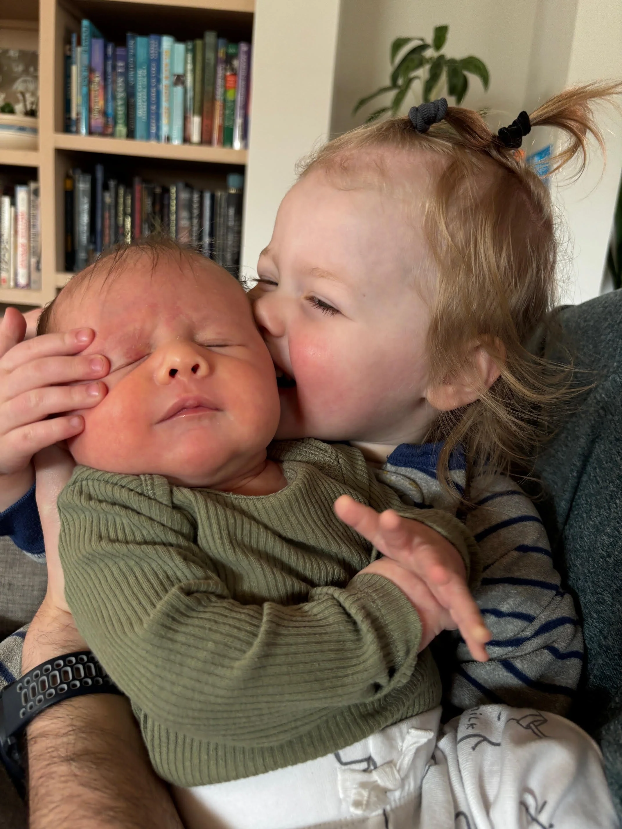 An older toddler with white skin and blond hair in two pigtails is kissing her younger sister, who is red in the face with eyes closed.