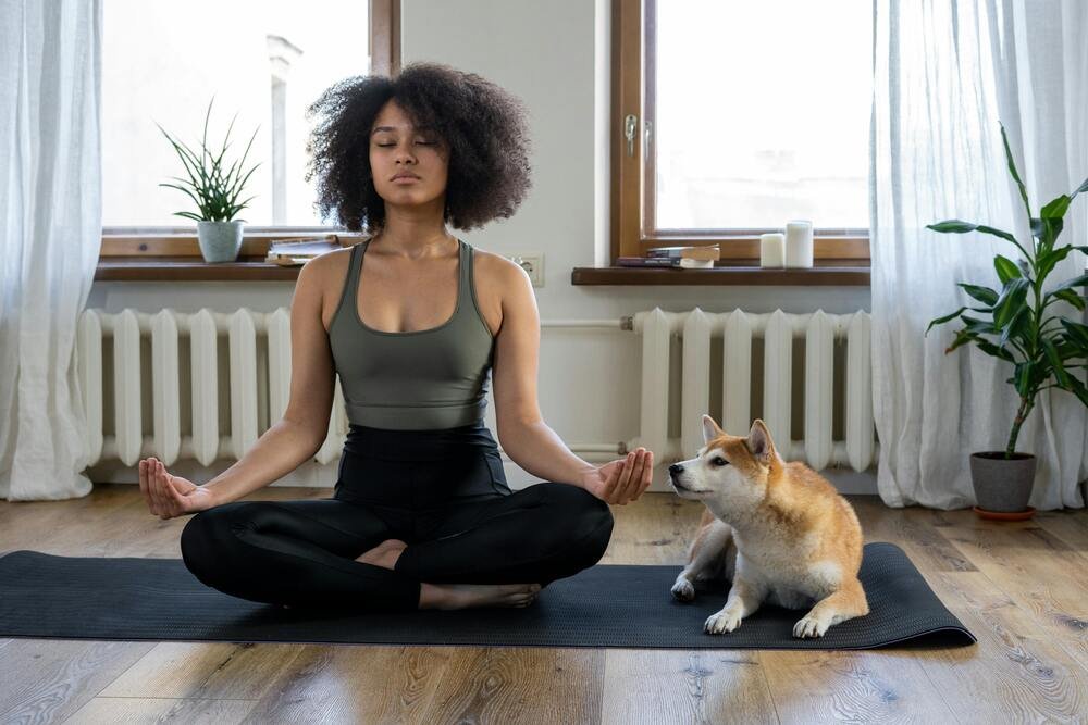 Woman in lotus position practicing self-care meditation indoors with her dog, promoting relaxation and calm in a peaceful home environment