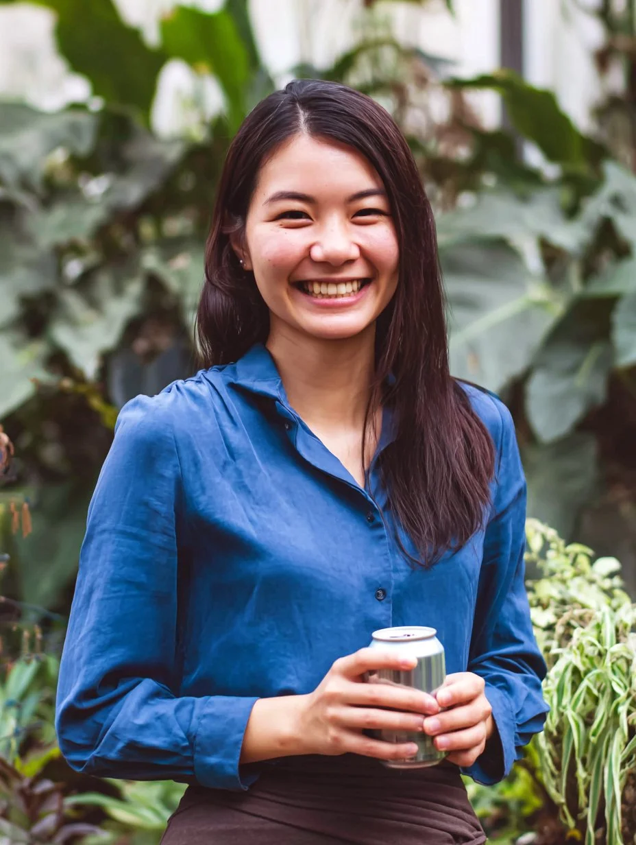 A woman smiling and holding a can inside a lush green greenhouse or botanical garden.
