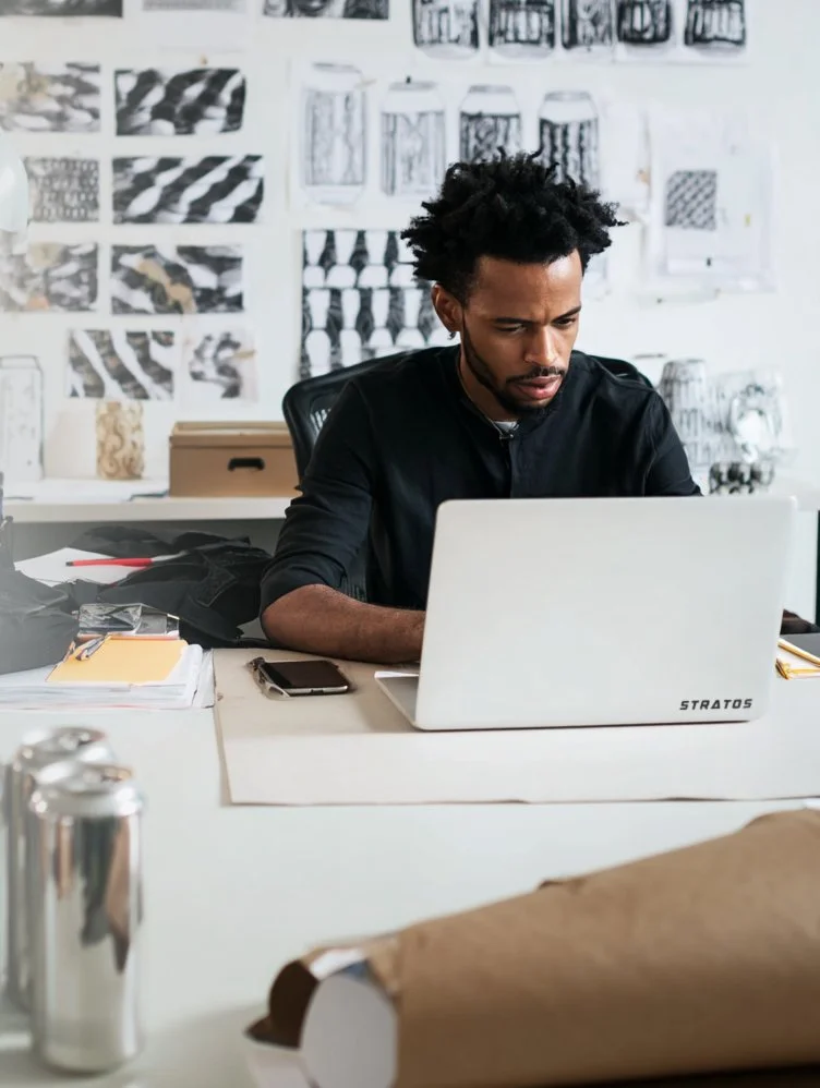 A man with curly hair and a beard working on a white laptop at a desk in an office. Art Director, creative type.
