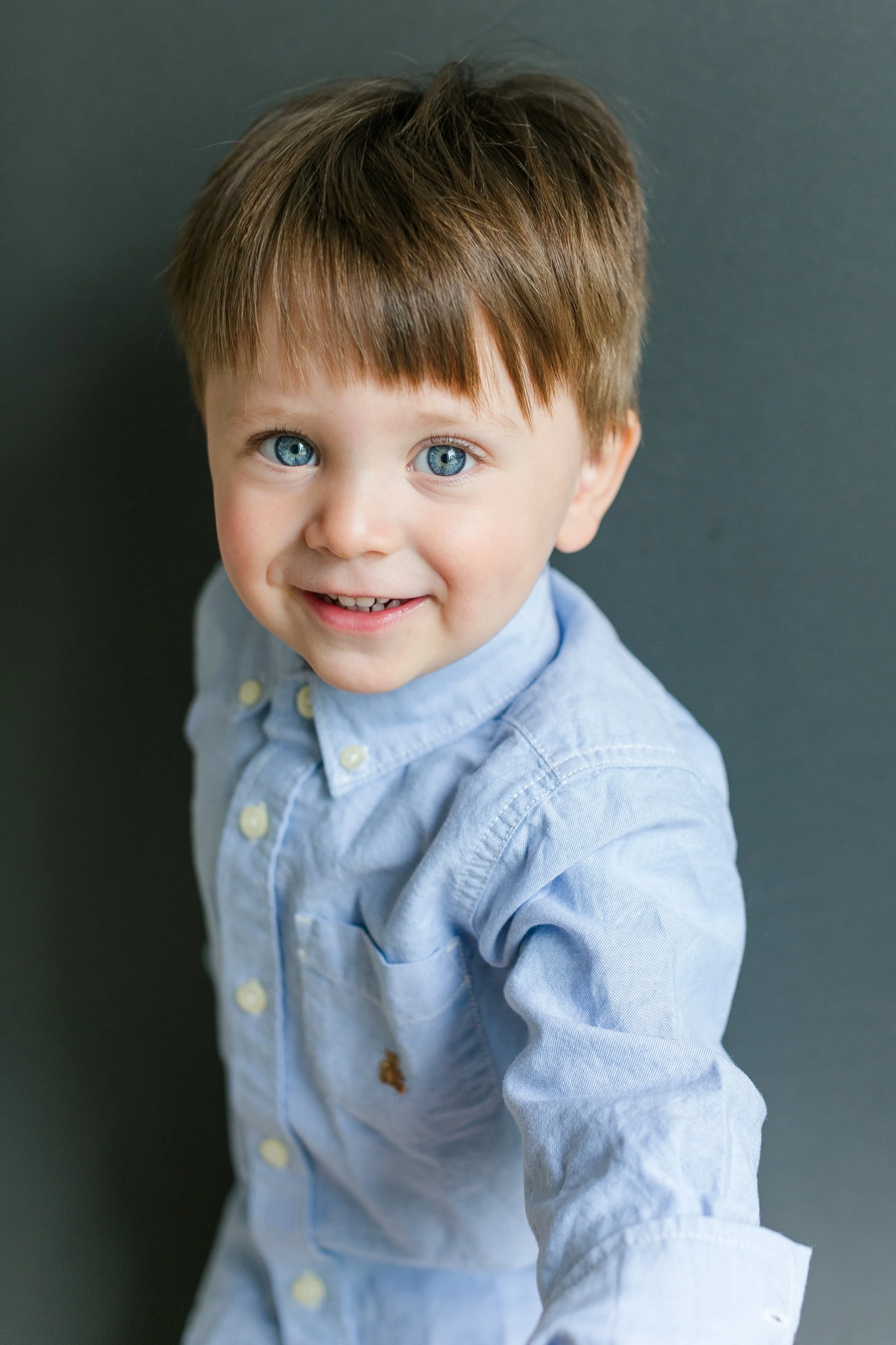 A young boy with blue eyes and light brown hair smiling at the camera, wearing a light blue button-up shirt, against a dark background.