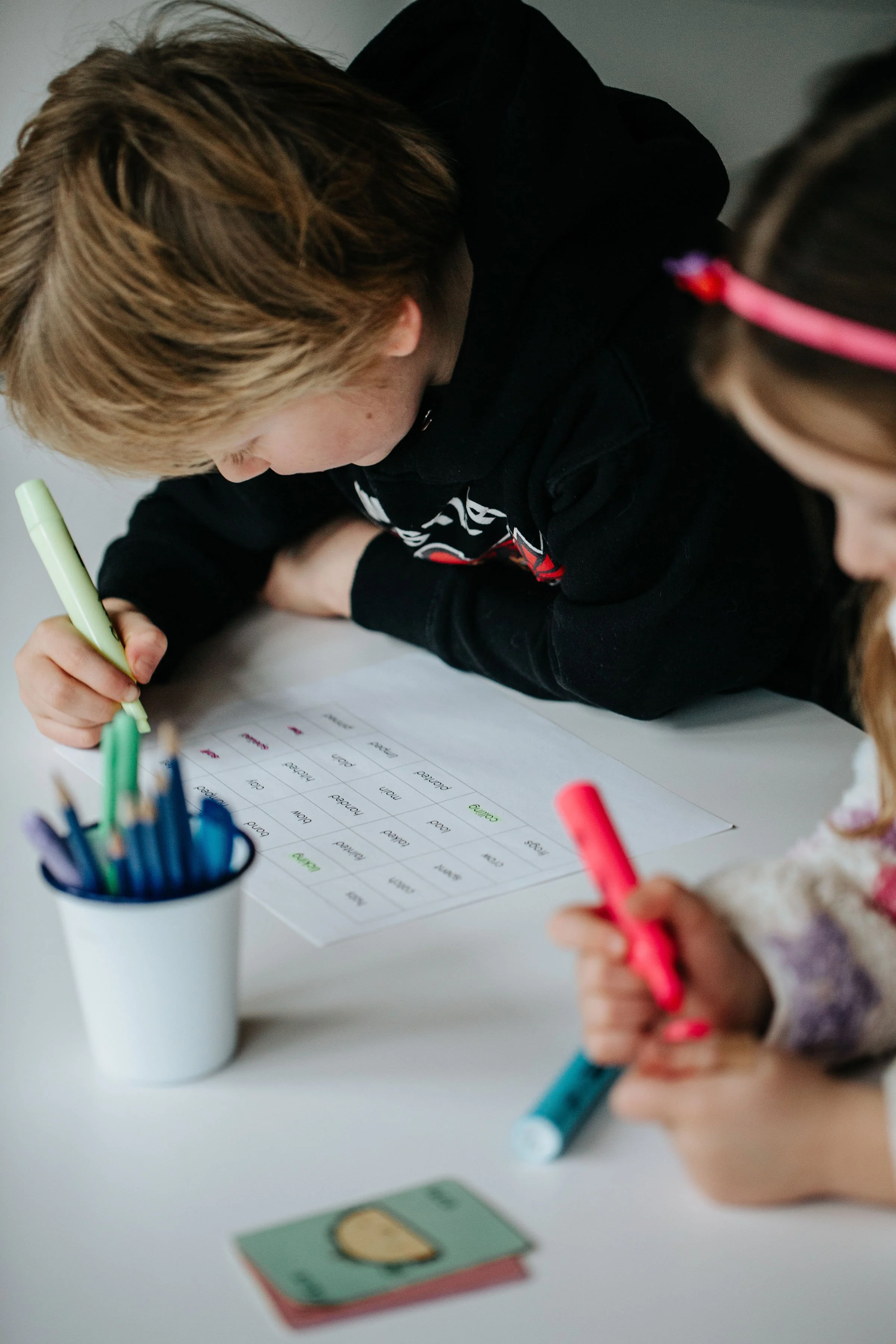 Two children practicing handwriting at a white table, with a worksheet, markers, and a cup of colored pens nearby.