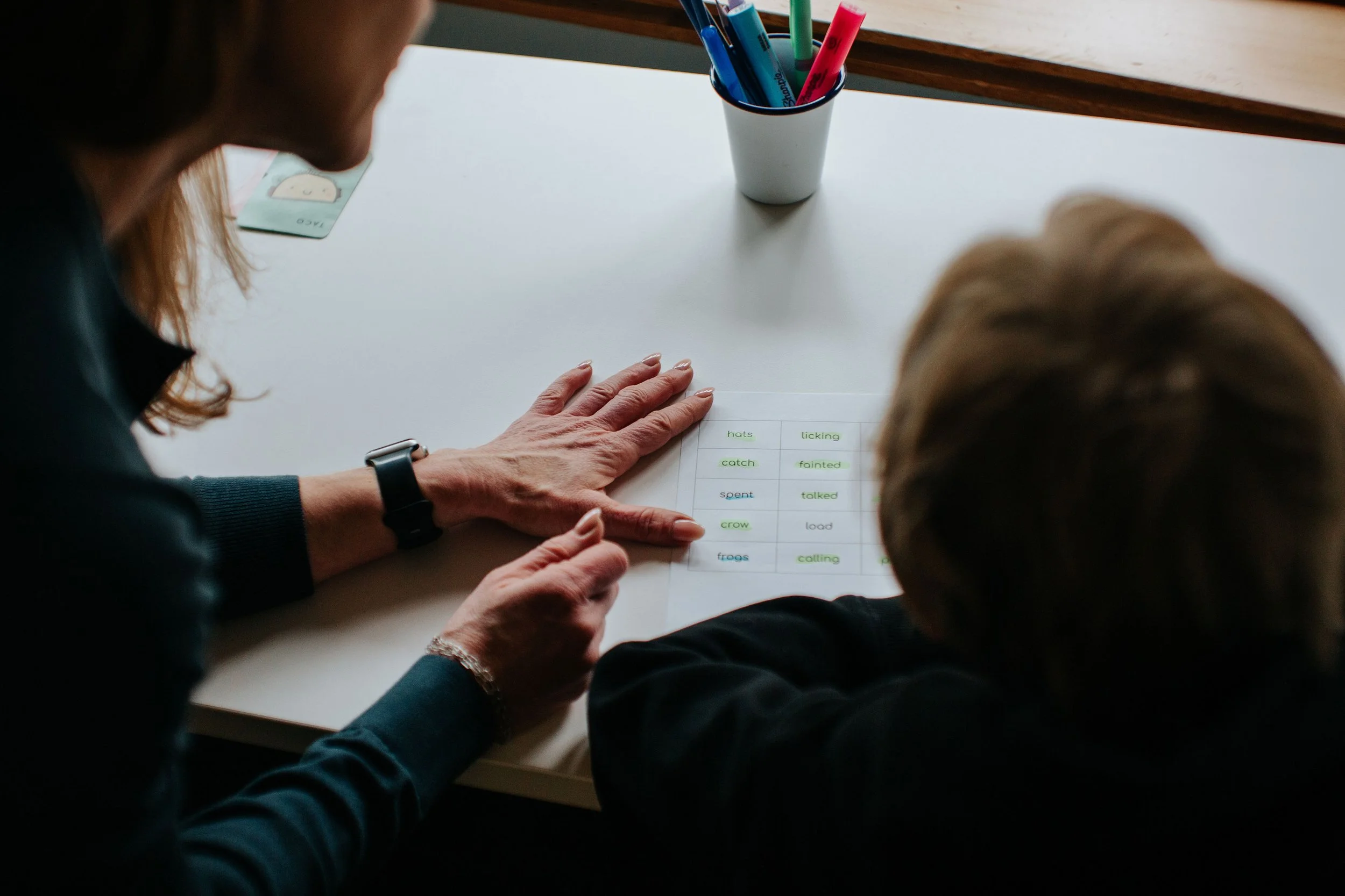 An adult and child looking at a sheet of paper with words in different colors, on a white table with a cup of markers and a card nearby.