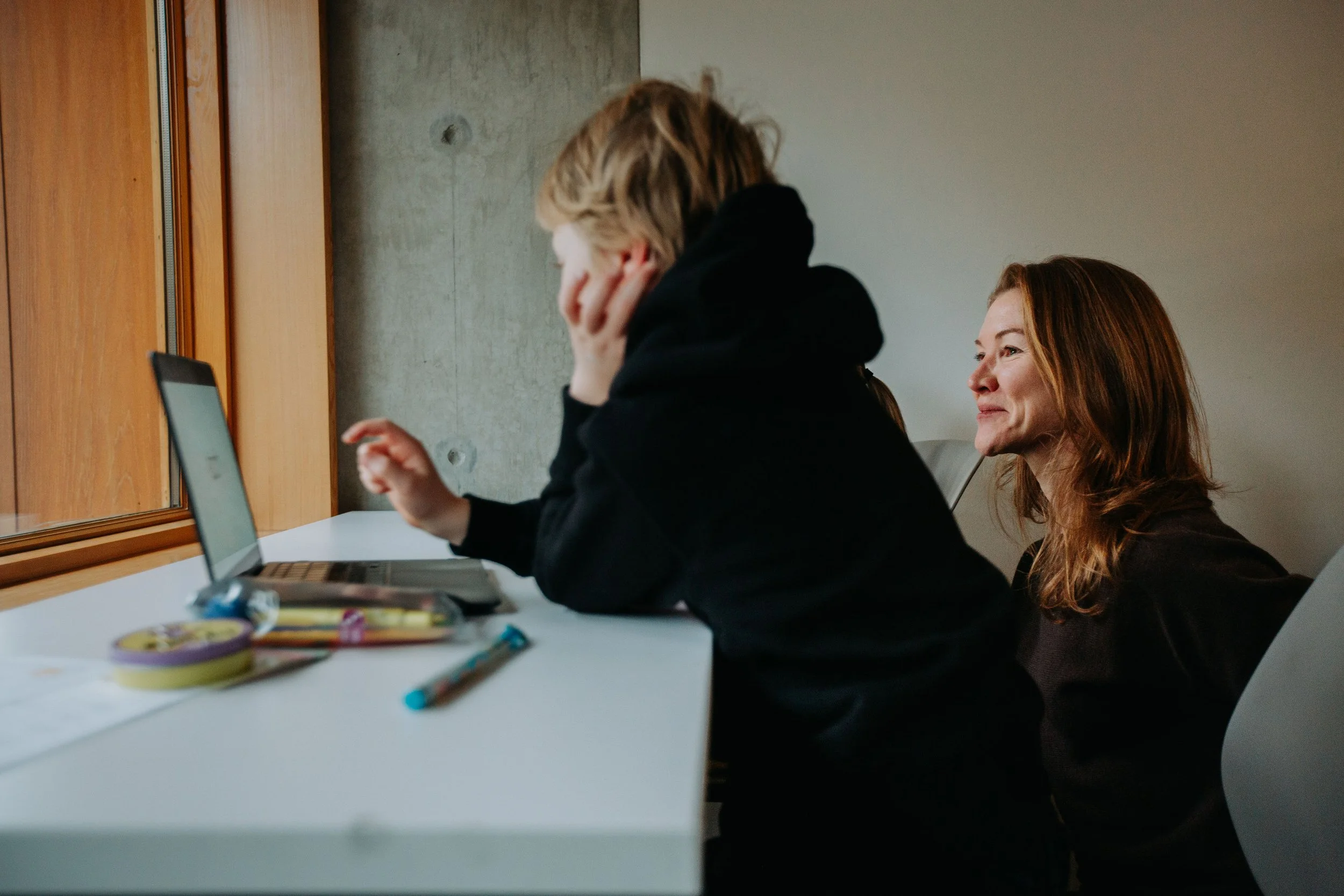 A woman smiling while sitting next to a child using a laptop at a table, with stationery items on the table and a window behind them.