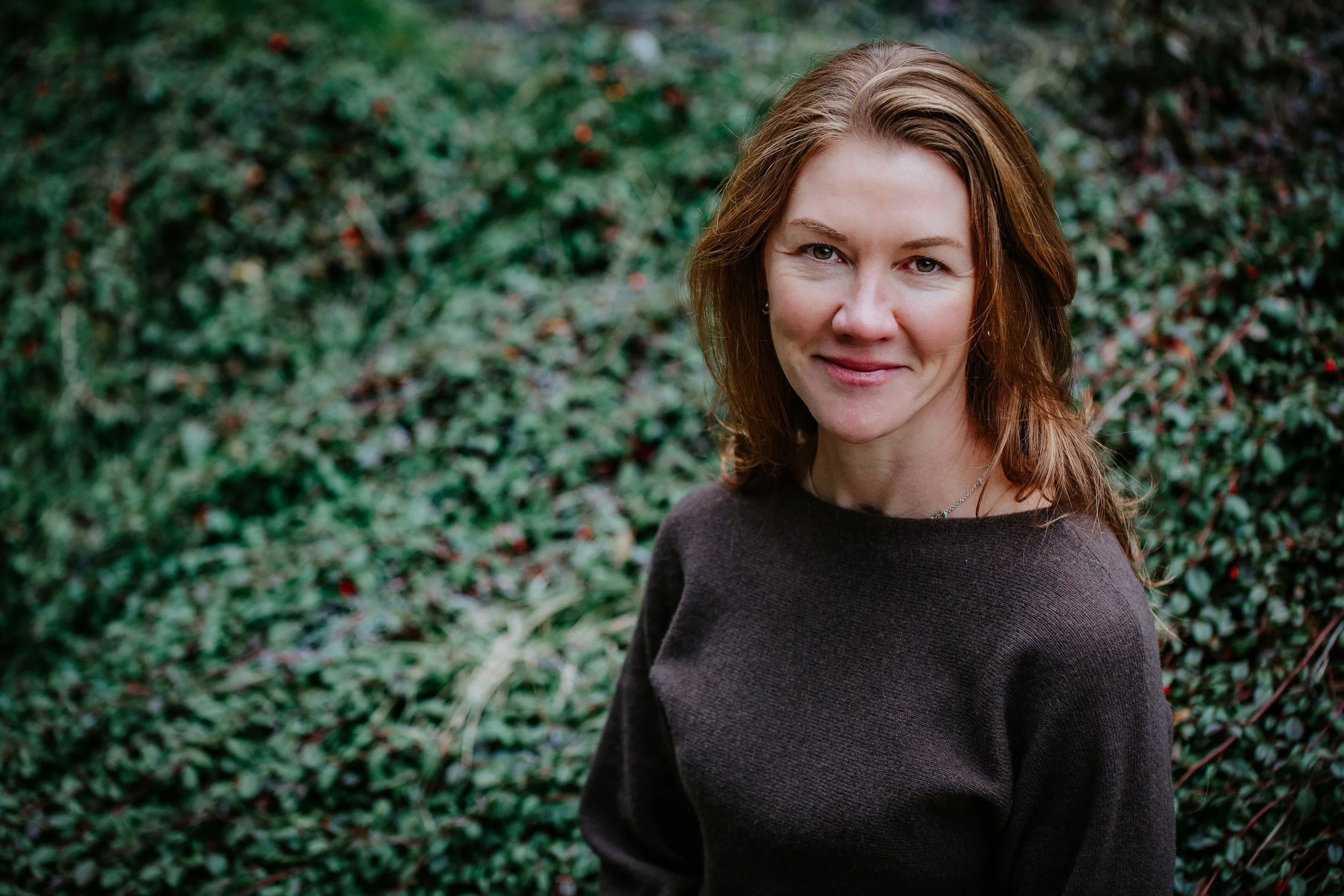 A woman with shoulder-length brown hair wearing a dark sweater, smiling outdoors with a background of green foliage.