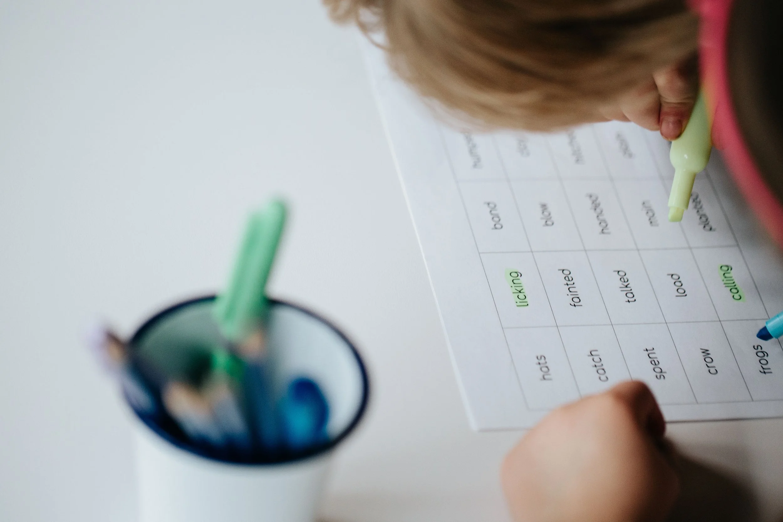 A child learning to read using a yellow marker to circle a word on a printed sight words list, with a cup of markers in the foreground.