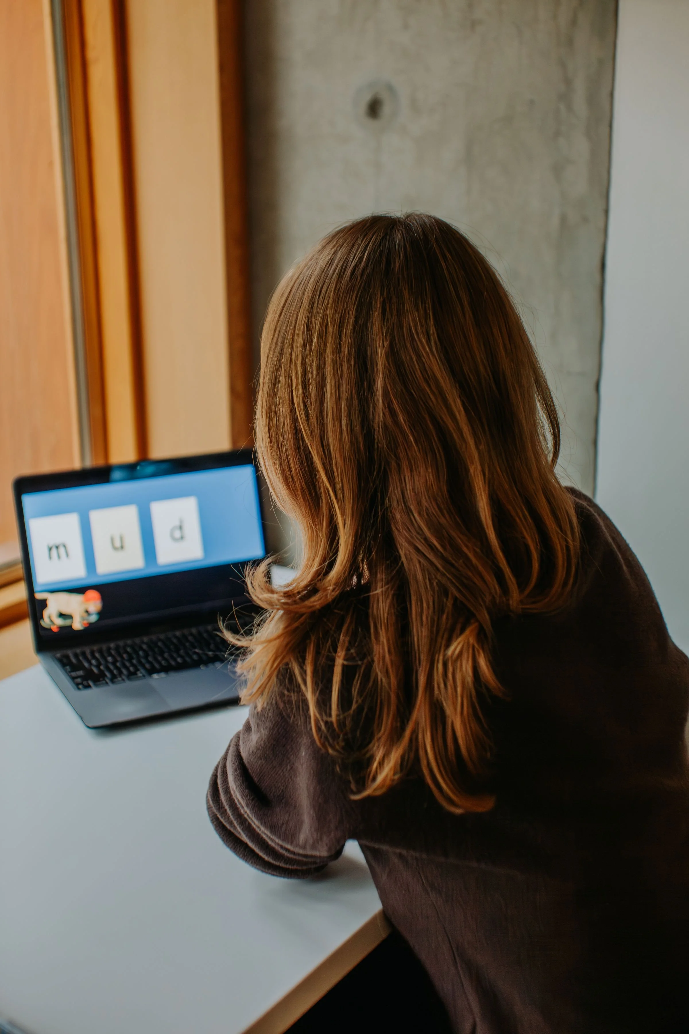 A person with long brown hair sitting at a desk, looking at a laptop screen displaying the word 'mud' with three letter tiles and a small cartoon lion in the bottom left corner.