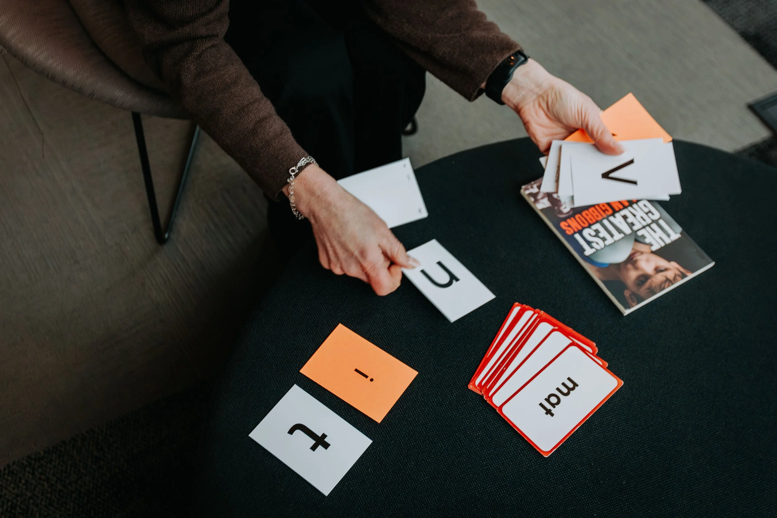 A person is sitting at a black table with reading cards and papers, including the title 'The Greatest in the Bluebonnets.' They are holding a plan 'u' card and there are other cards and papers scattered on the table.