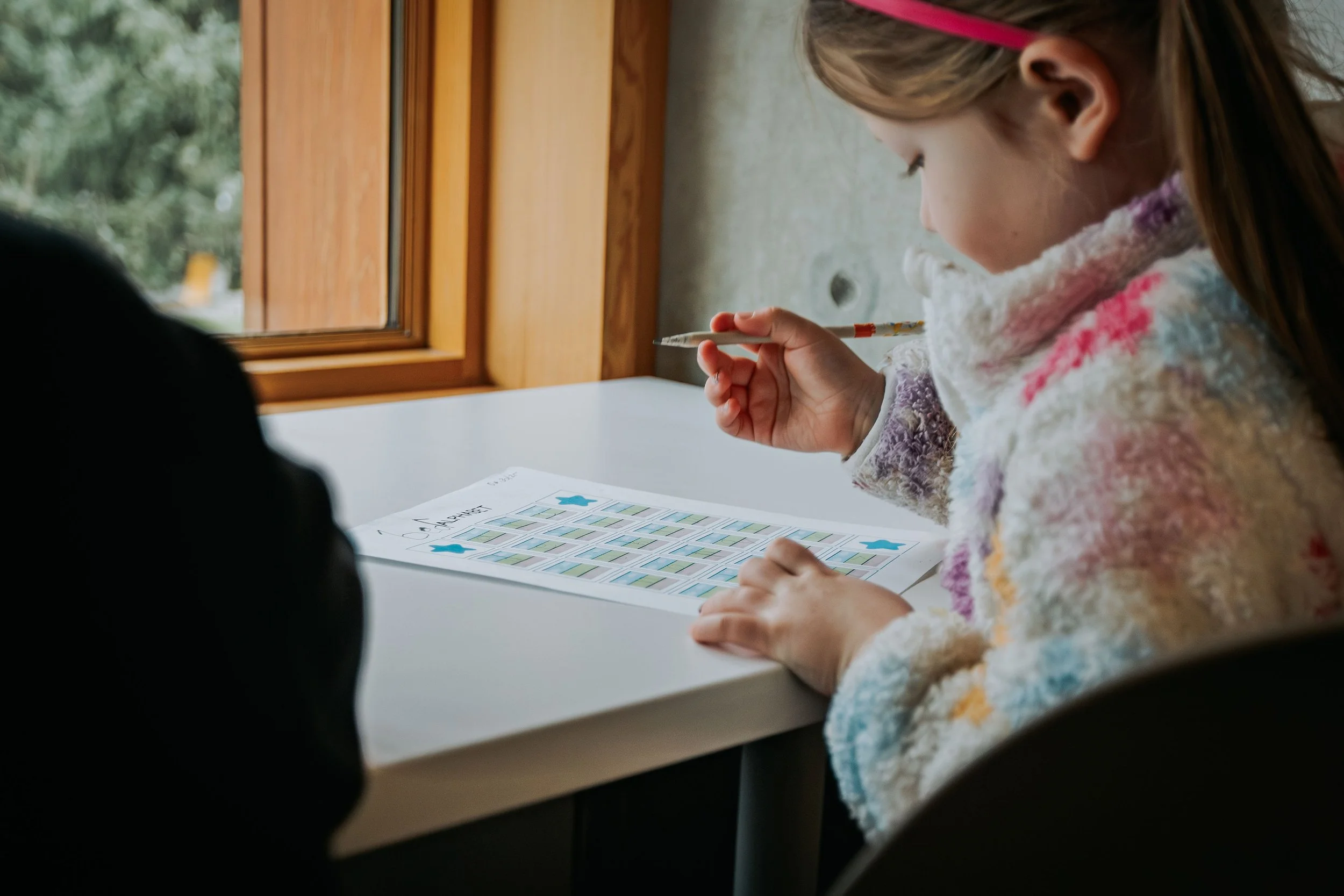 A girl with a colorful sweater sitting at a white table, looking at a printed sheet with literacy exercises featuring blue star stickers, holding a pen near her face, near a window.