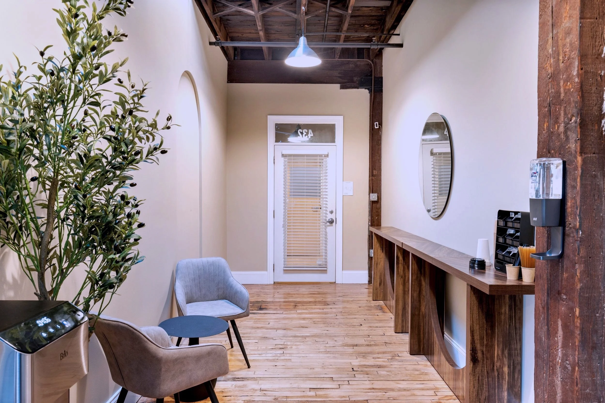 HWHC Minimalist waiting area with two gray chairs, a round table, an indoor plant, a wooden counter, a water dispenser, and a door with blinds.