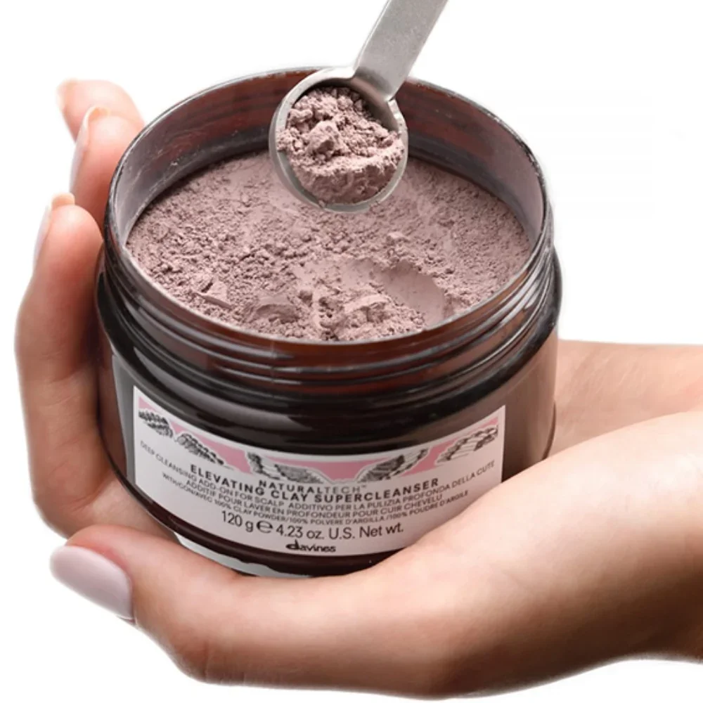Hand holding a jar of loose pink clay powder with a metal scoop.