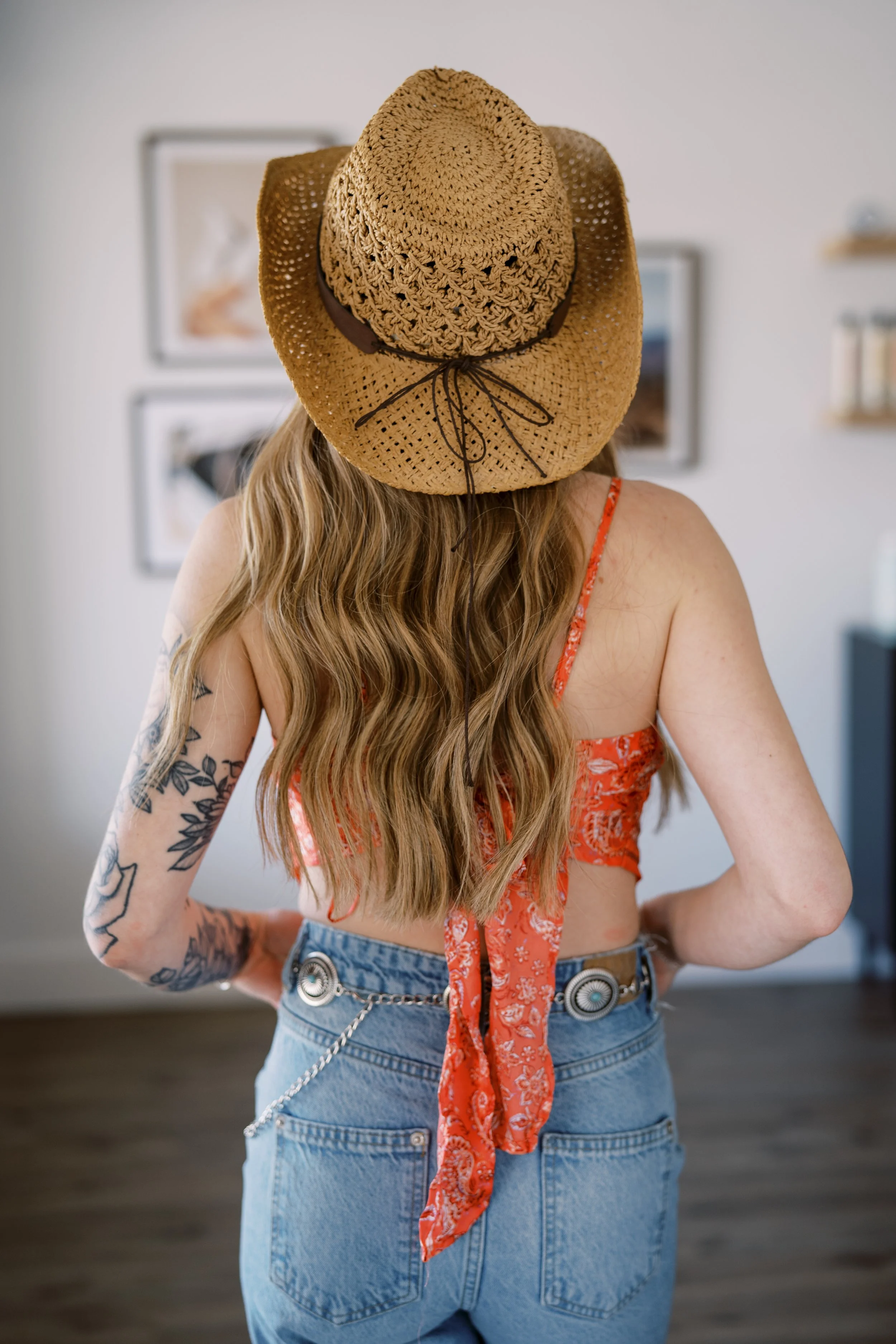A woman with long wavy hair wearing a straw sun hat, red floral top with a tie at the back, and high-waisted light blue jeans with a chain and concho belt, standing indoors with artwork and shelves on the wall behind her.