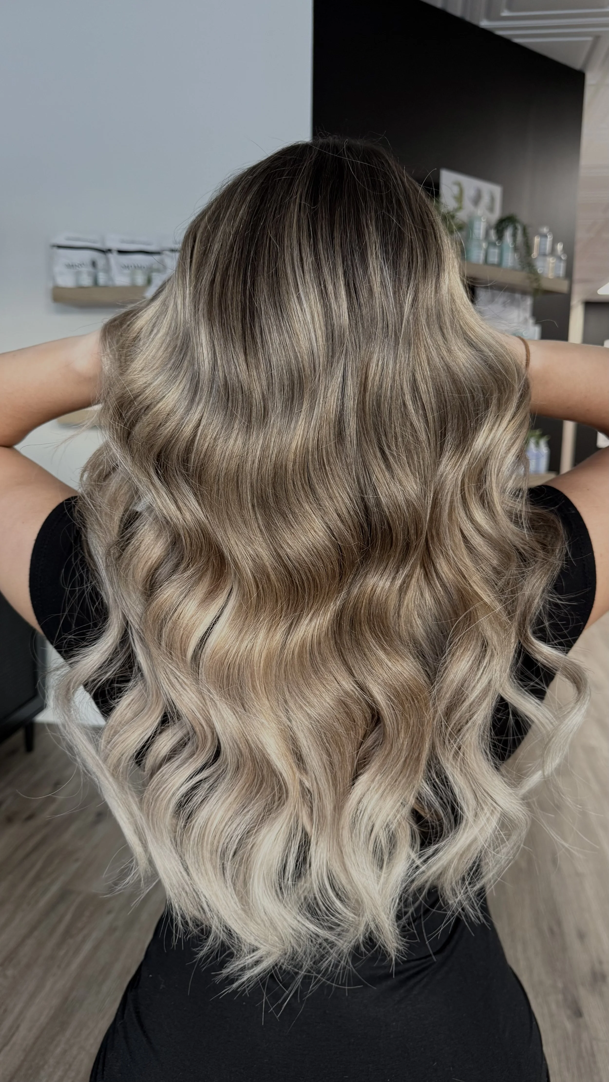 Back view of a woman with long, wavy hair dyed in a gradient from dark brown at the roots to blonde at the ends, posing with her arms raised behind her head in a salon setting.