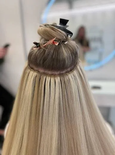 A woman at a hair salon is getting her hair dyed or styled. The top section of her hair is wrapped in foil, and she has a small black hat and a hair clip on top of her head. Her long, straight blonde hair is visible below.