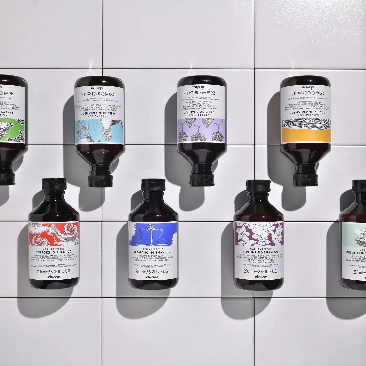 Multiple bottles of hair care products, including shampoos with labels displaying artistic designs, placed on a white tiled background.