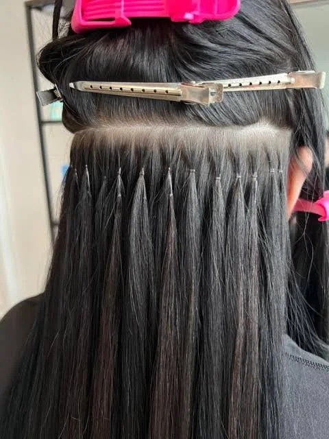 A person undergoing hair extension process, with hair sectioned and clipped back using pink and silver clips, and hair strands secured on a horizontal tape.