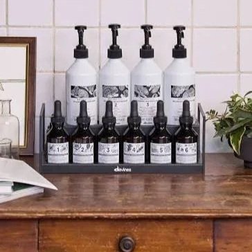 Two rows of black dropper bottles on a black rack on a wooden surface, with larger white bottles behind, set against a tiled wall with a plant to the right.