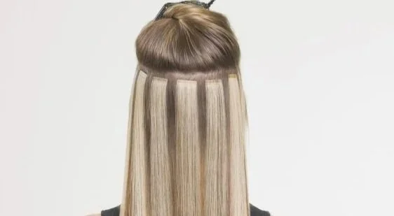 Hair extensions being attached to a person's straight blonde hair in a salon setting.