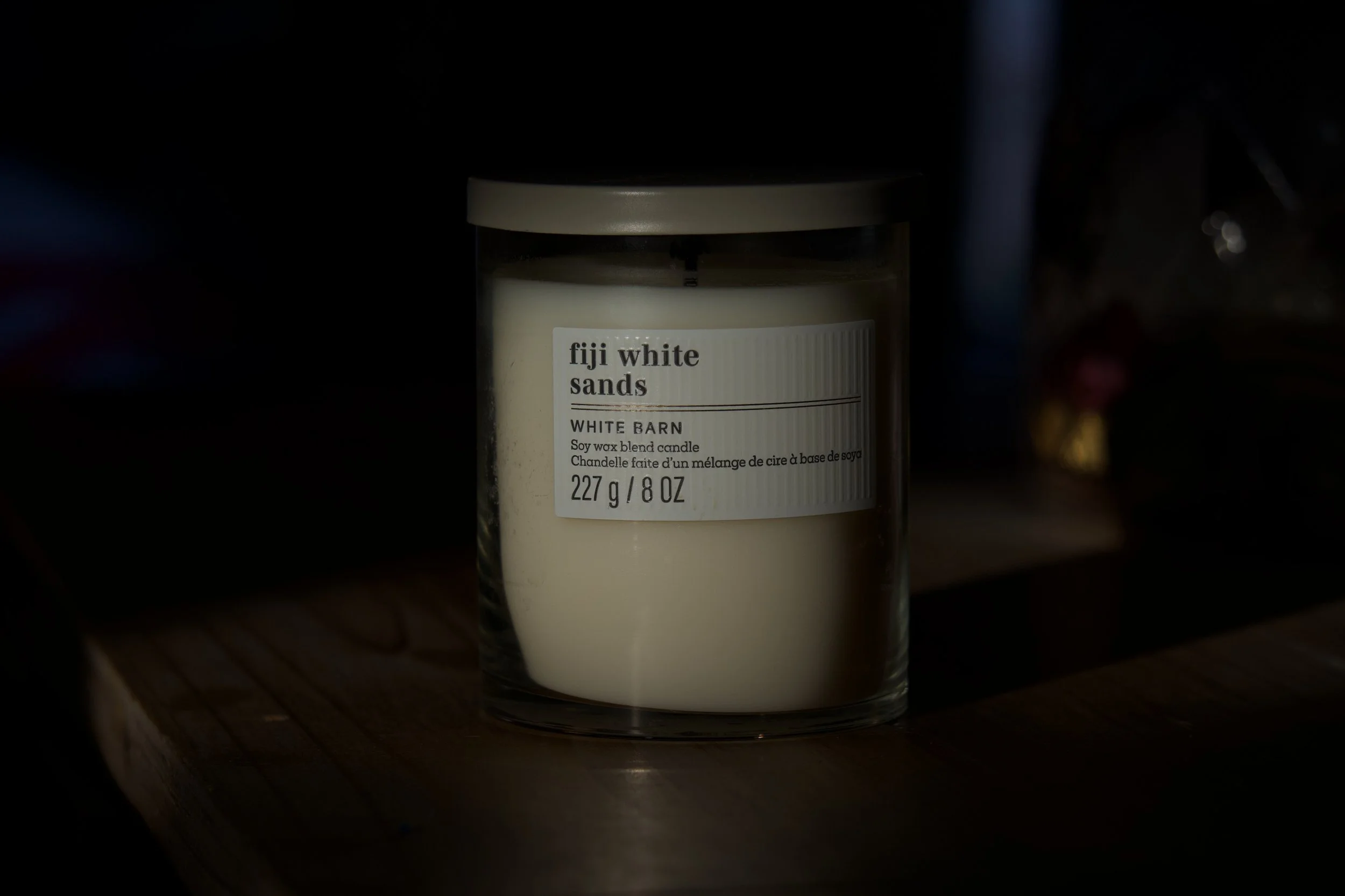 A white candle labeled 'fiji white sands' in a glass jar, placed on a wooden surface with dark surroundings.