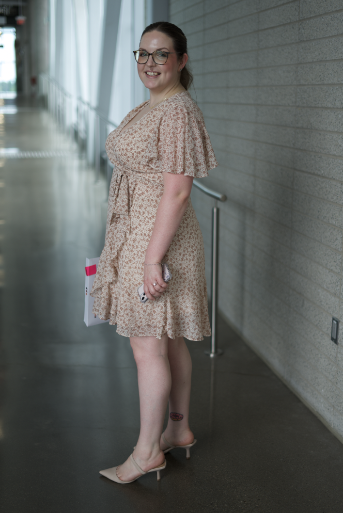 A woman in a beige floral dress and high heels standing inside a modern building with large windows and a textured wall, holding a phone and a folder, smiling at the camera.