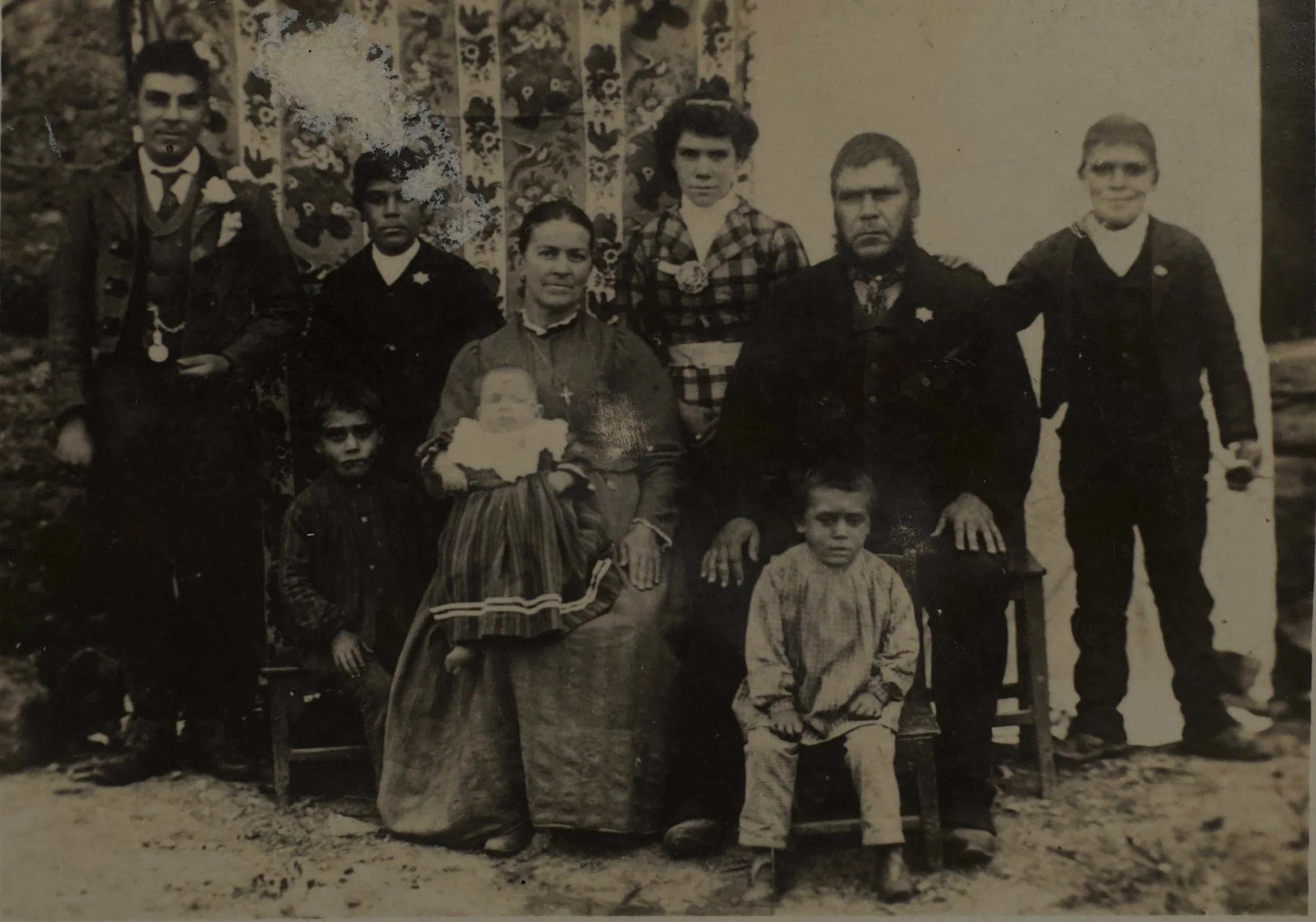 Black and white vintage family portrait of six children and a woman posed outdoors, with some children sitting and others standing. A floral curtain is in the background.