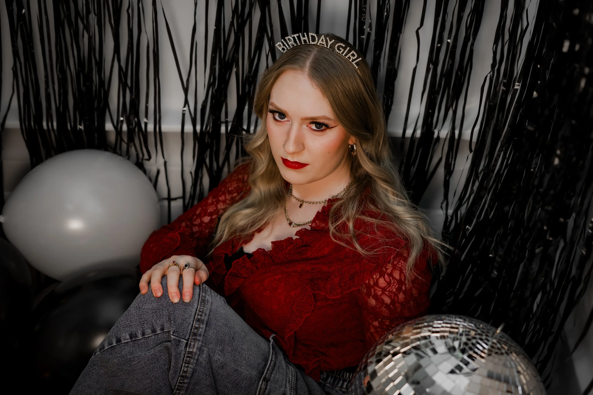 A woman with long blonde hair and red lipstick wearing a 'Birthday Girl' headband, red lace top, and jewelry, sitting among black and silver birthday decorations and balloons.