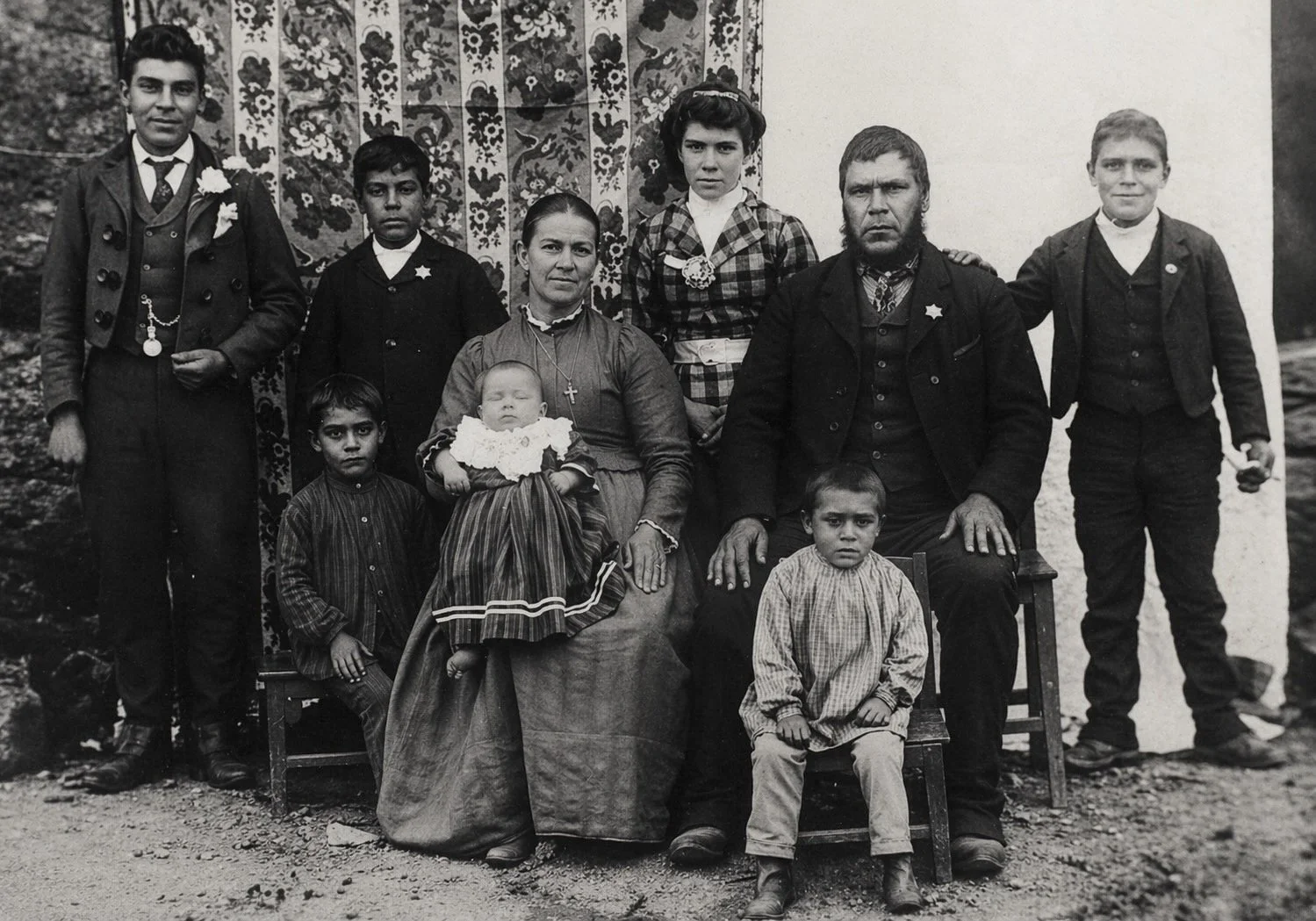 A black and white vintage photo of a family portrait outdoors, with nine people including men, women, and children, some dressed formally, and a decorative fabric backdrop behind them.