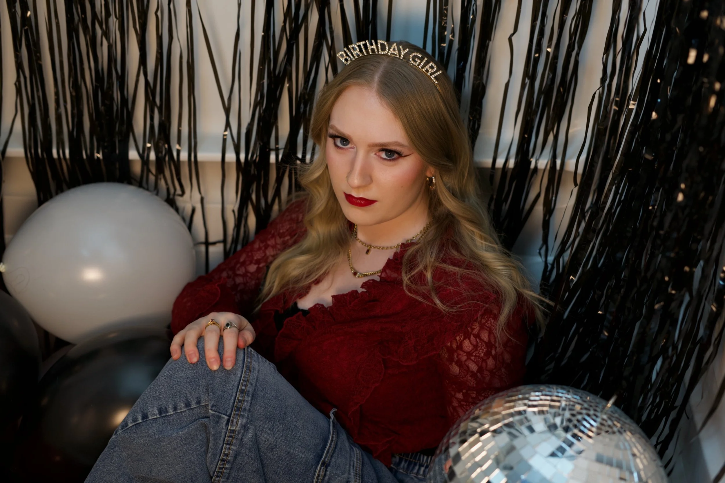 A young woman with blonde hair and red lipstick sitting inside a black fringe backdrop with balloons, wearing a 'Birthday Girl' headband, red lace top, and multiple necklaces, with a shiny disco ball in the foreground.