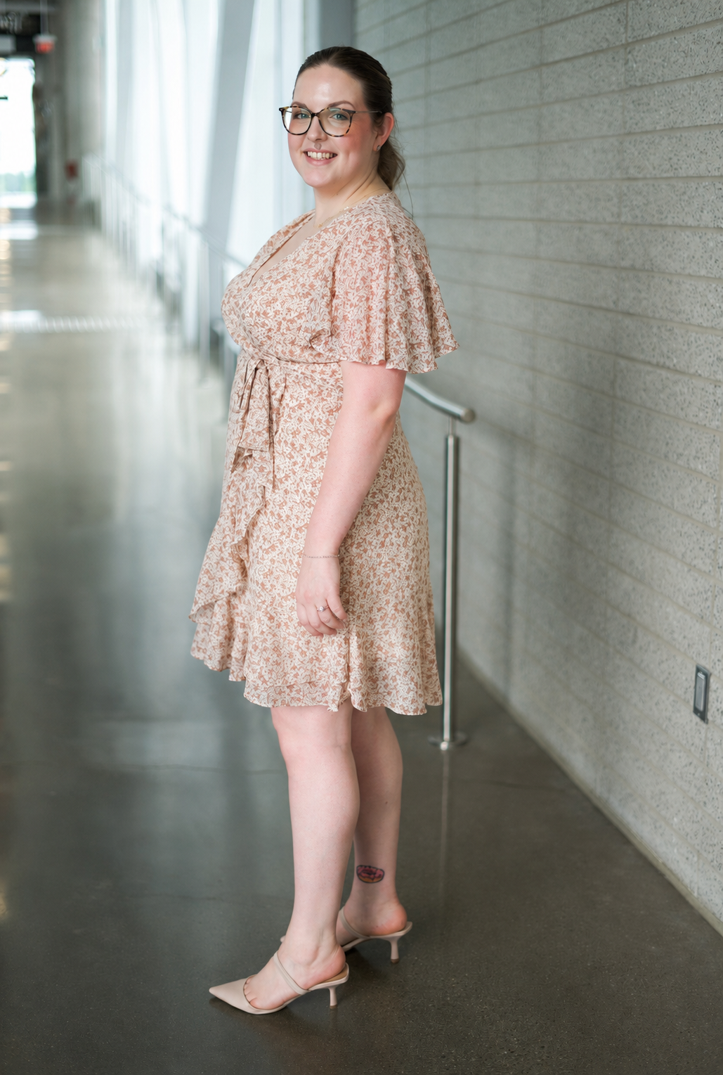 A woman with glasses and a tattoo on her ankle smiling while standing in a hallway, wearing a peach-colored floral dress and nude heels.