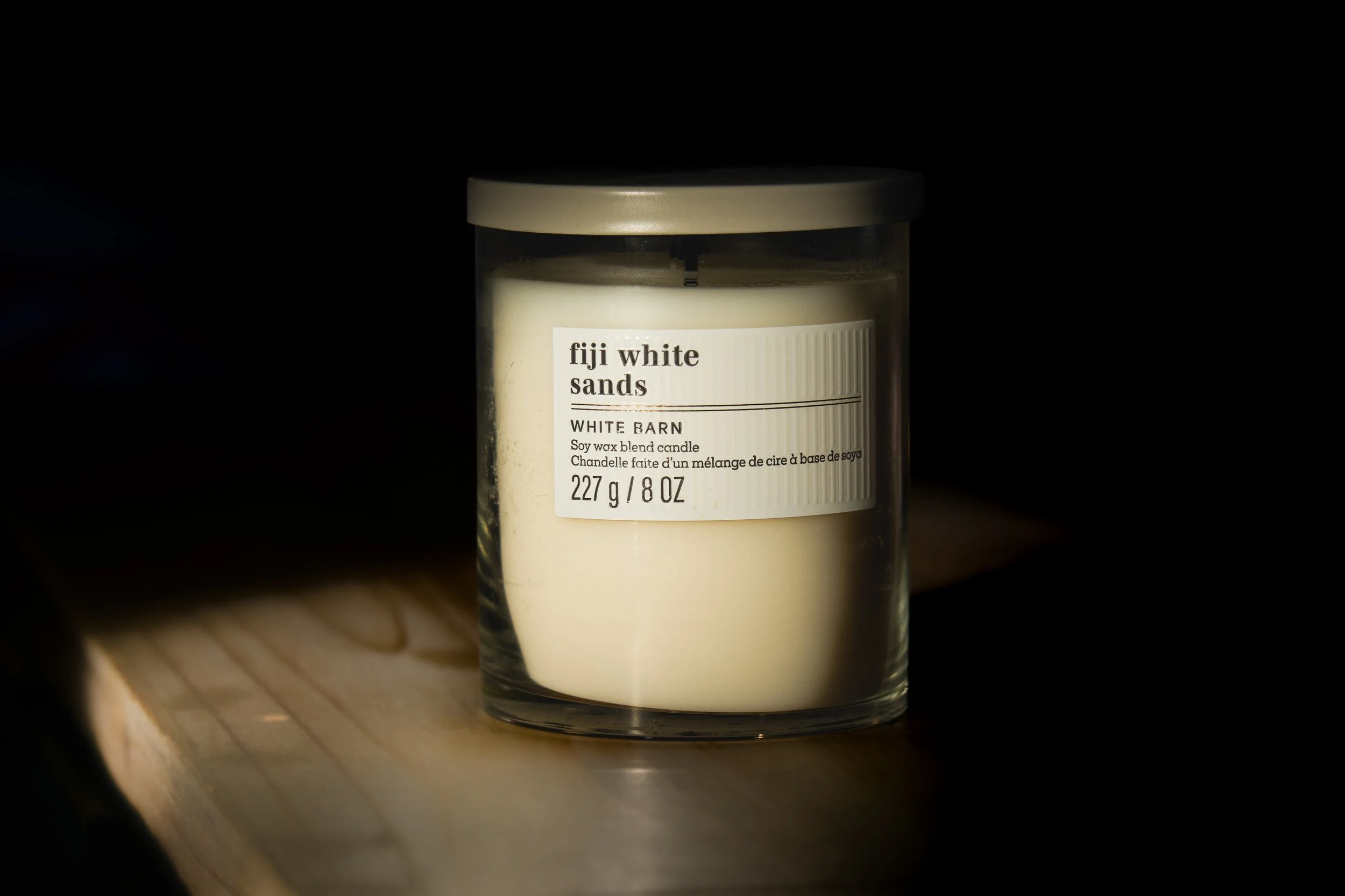 A white barn scented soy wax candle in a clear glass jar with a metal lid, placed on a wooden surface with a dark background.