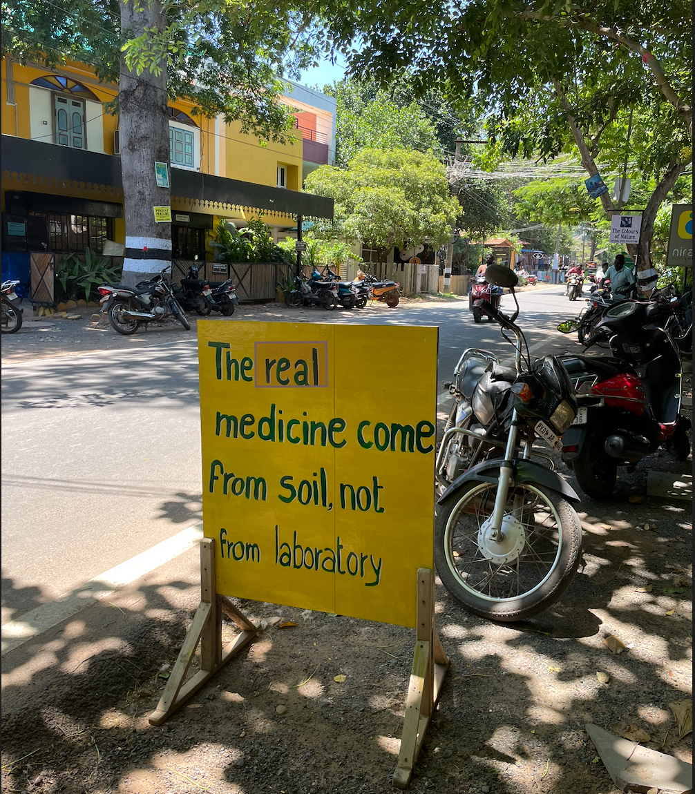 Yellow signboard on a street corner with green text reading 'The real medicine come from soil, not from laboratory,' next to a parked motorcycle. Background shows trees, parked scooters, and colorful buildings on a sunny day.