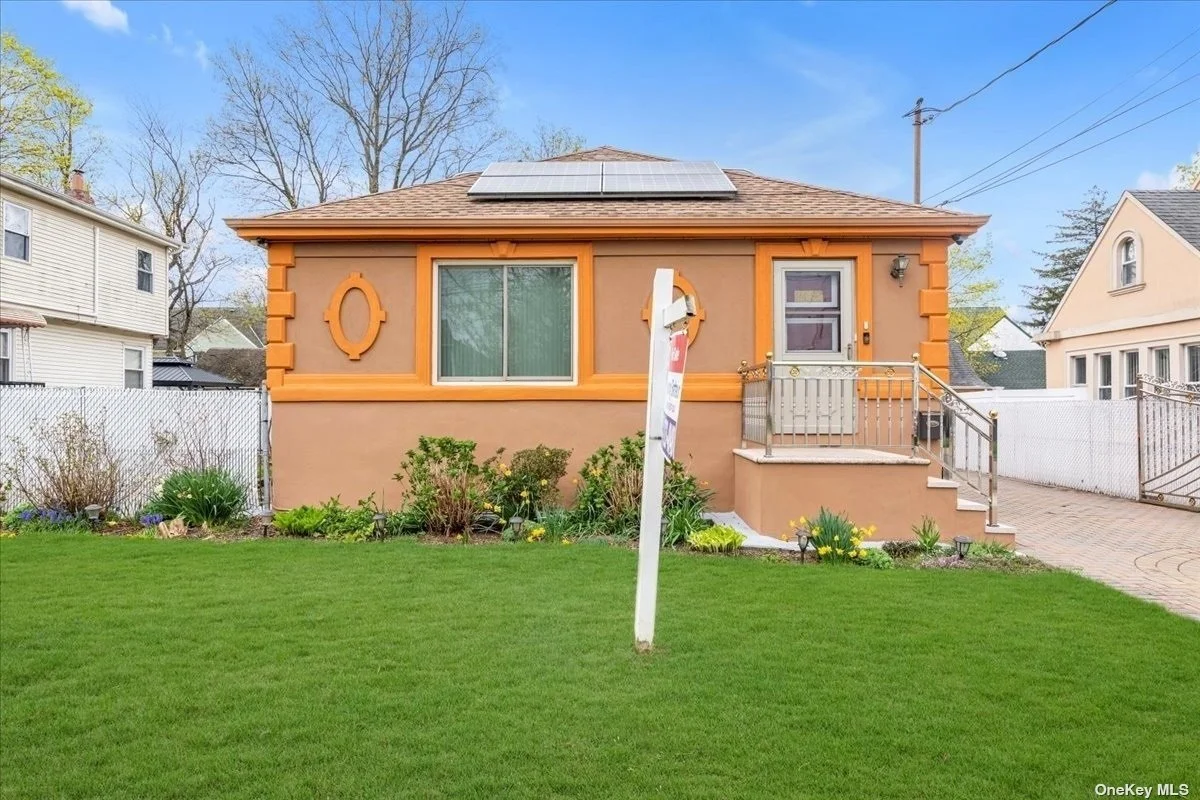 A small house with orange trim and a solar panel on the roof, front yard with green grass, plants, and flowers, white picket fence on sides, walkway leading up to the front porch, window with curtains, and porch with steps and railing.