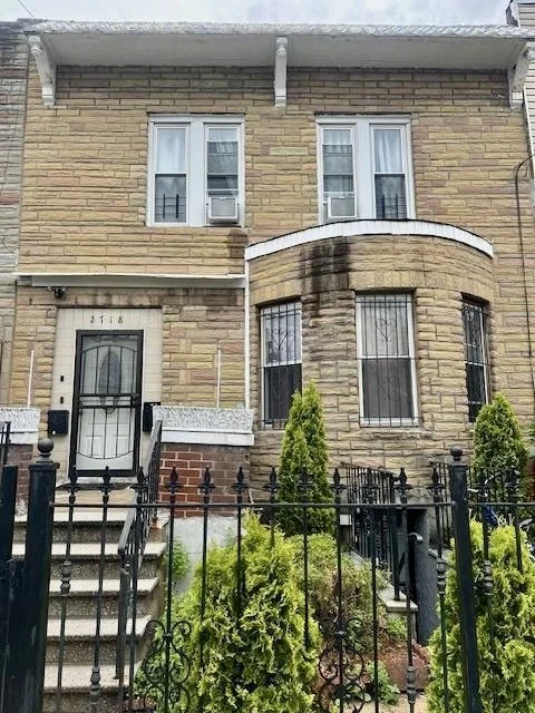 Front view of a two-story brick house with a black iron fence and small garden with shrubs. The house has a bay window, four windows on the upper floor, and a front door with steps.