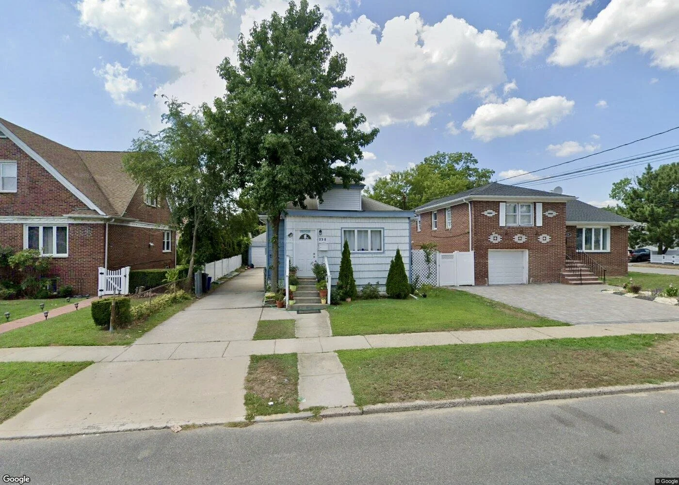 A suburban street view with a white house in the center, flanked by a red brick house on the left and a modern multi-story brick house with a garage on the right. There are trees, a sidewalk, and a paved driveway.