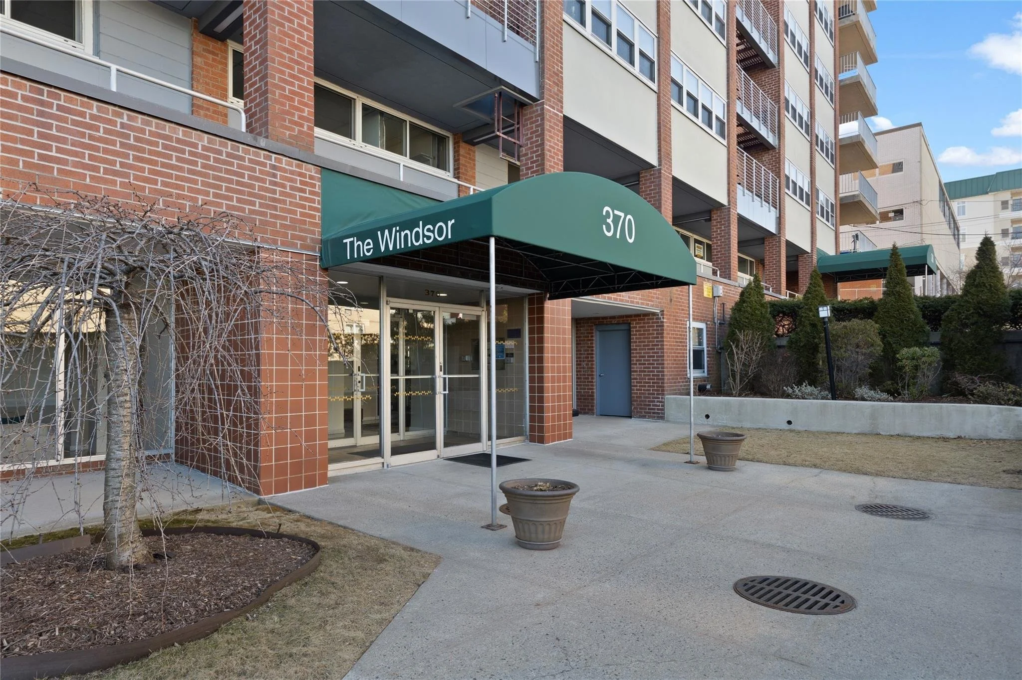 Apartment building entrance with green awning labeled 'The Windsor' and address number 370, glass doors, planters, and landscaped area with bushes and small trees.