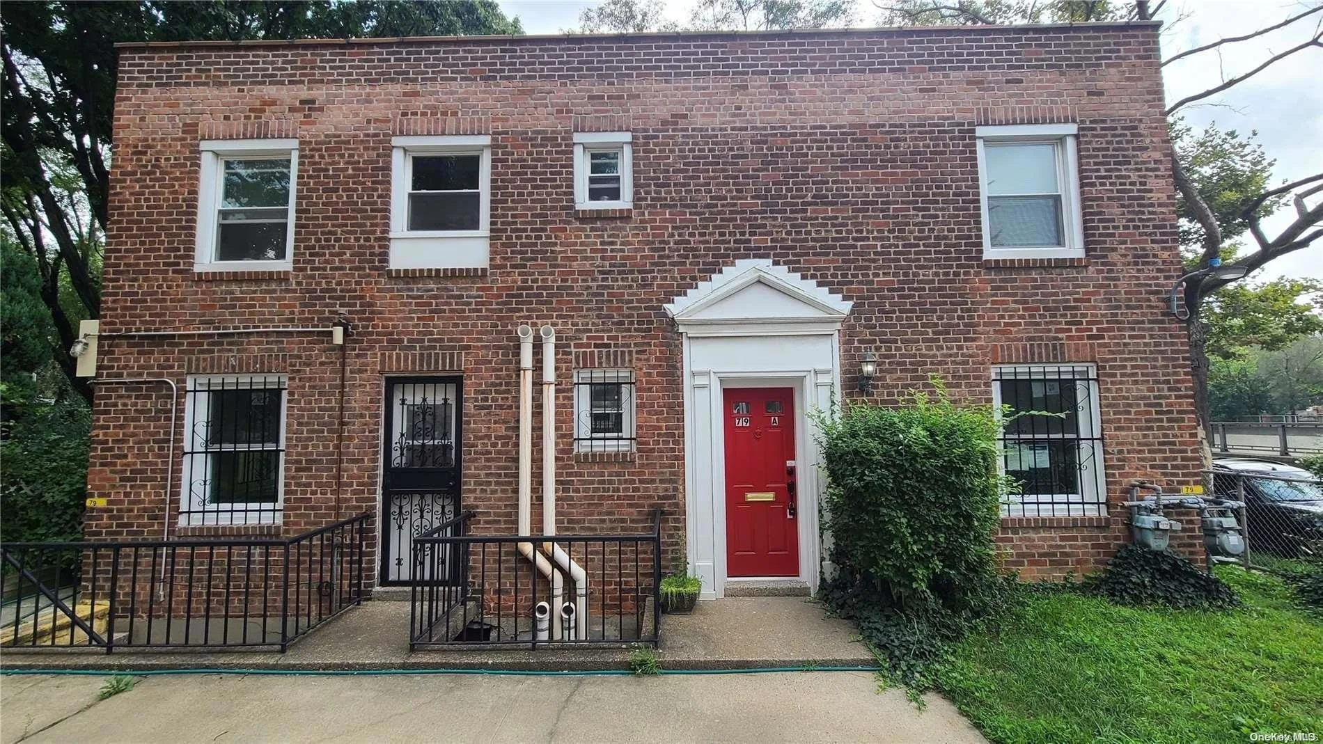 Two-story brick residential building with a red front door, three second-floor windows, two first-floor windows with iron bars, a small gate on the left, some plants and bushes near the entrance, and utility meters on the right side.