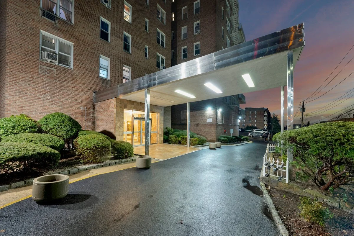 Apartment building entrance with a covered drop-off area, surrounded by trimmed bushes, during dusk with a colorful sky.