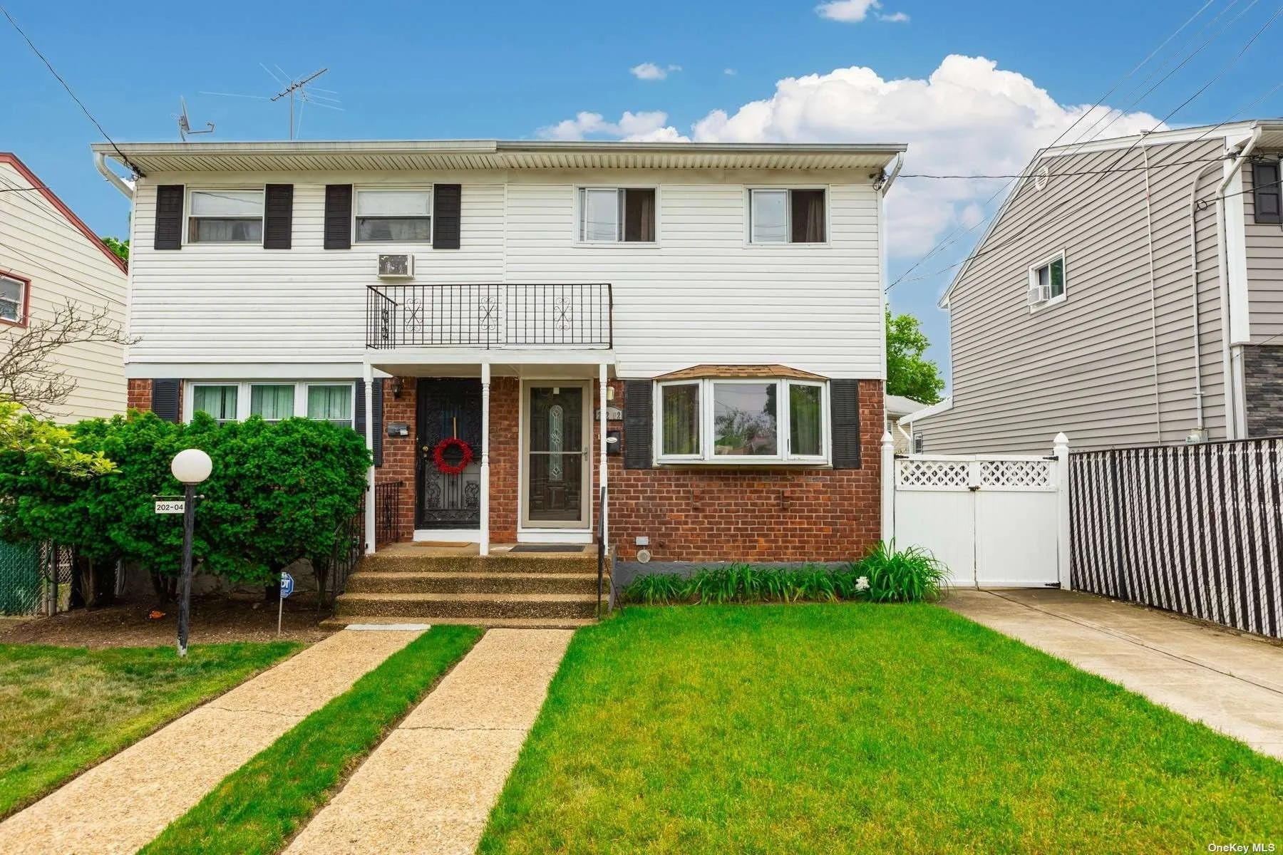 A two-story house with white siding, black shutters, and a brick lower facade. The house has a front door with a red holiday wreath, a small staircase, and a lawn with green grass and a small bush. There is a sidewalk and a lamp post near the stairs, and neighboring houses are visible on each side. The sky is blue with some clouds.