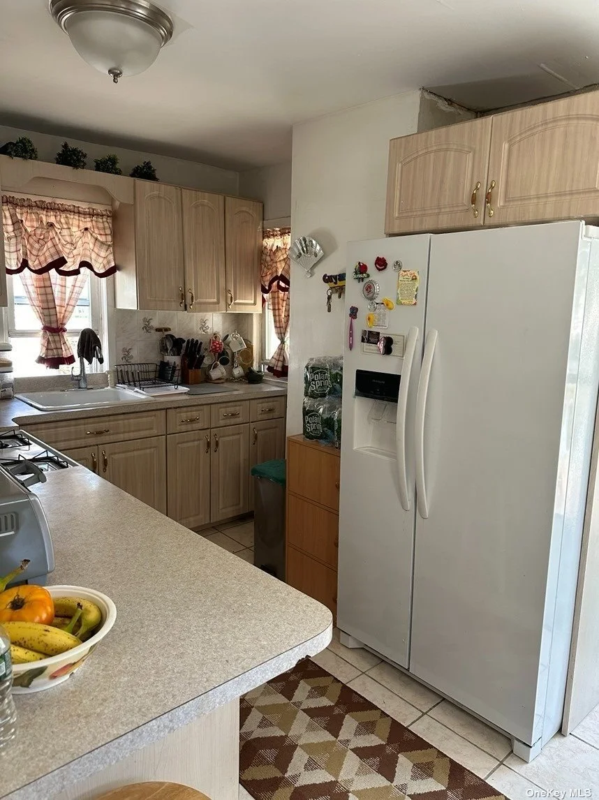 A kitchen with wooden cabinets, a white refrigerator with magnets, a countertop with a fruit bowl, and windows with red checkered curtains.