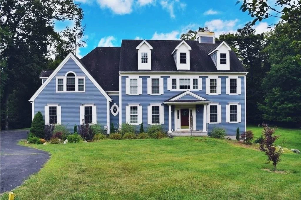 A large blue house with white trim, multiple gables, and a front porch, surrounded by a well-kept lawn and trees.