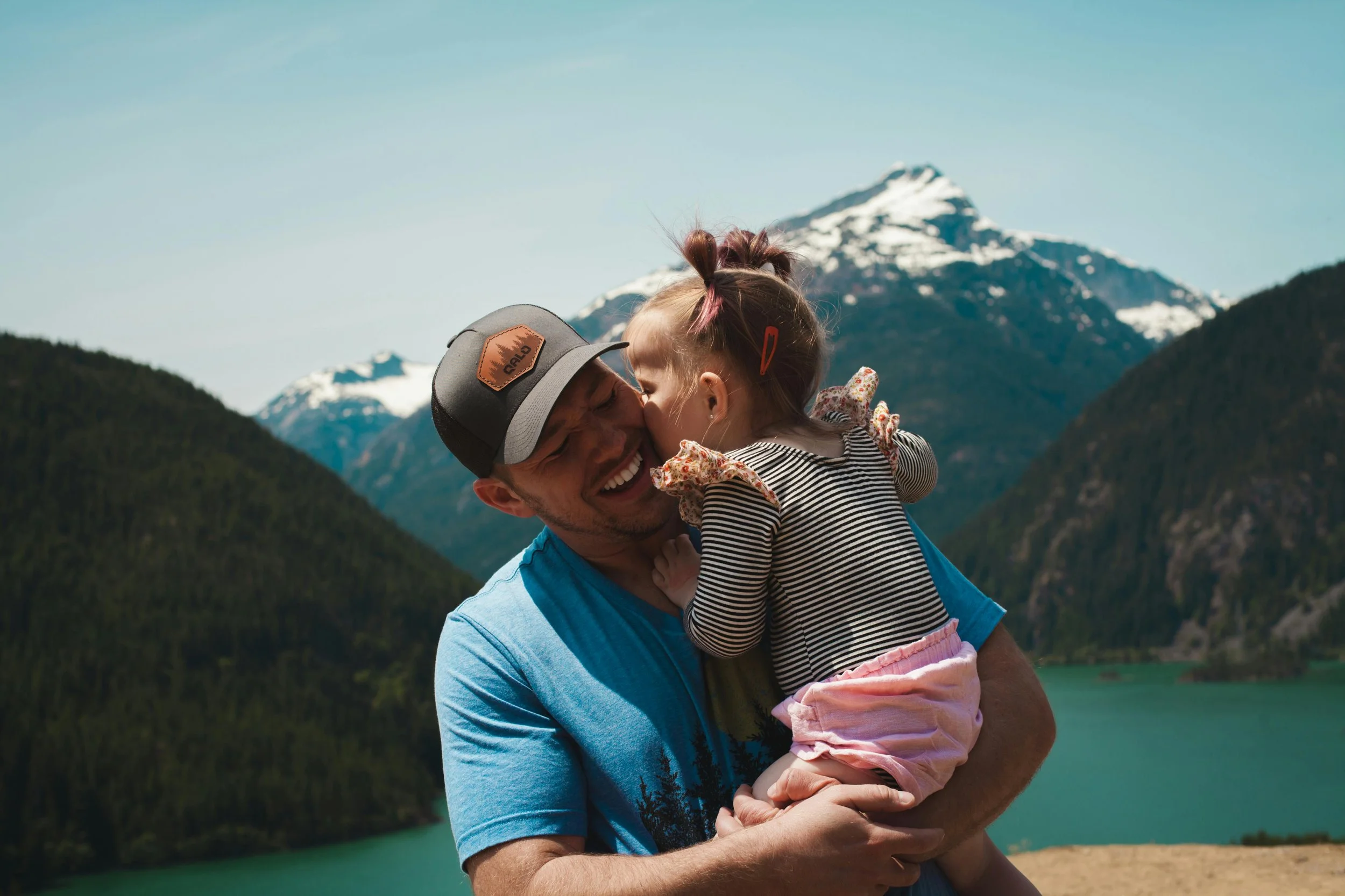 "Father holding and kissing his young daughter in front of a mountain lake, symbolizing the emotional journey of becoming a parent."